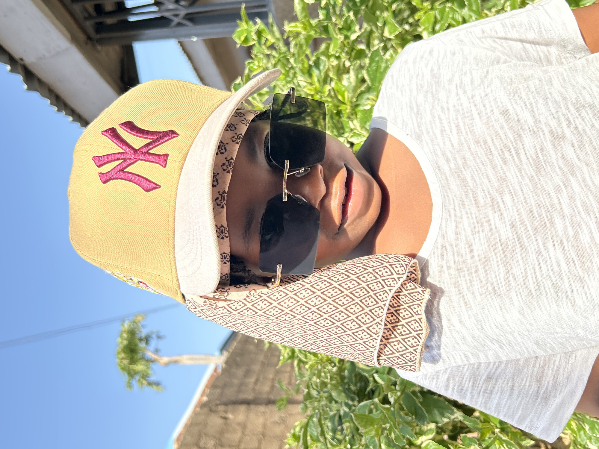 A person wears a tan baseball cap with a pink "NY" logo and a white brim. The cap has a patterned band below the brim. They are wearing square, black sunglasses with gold nose pieces. The person also has on a light-colored t-shirt. The picture was taken outside, with green plants in the background and a blue sky in the upper left corner. Part of a building structure is visible above the person's cap on the left side. While the specific location within Jos, Nigeria isn't evident, the lighting and environment suggest an outdoor setting in the city.