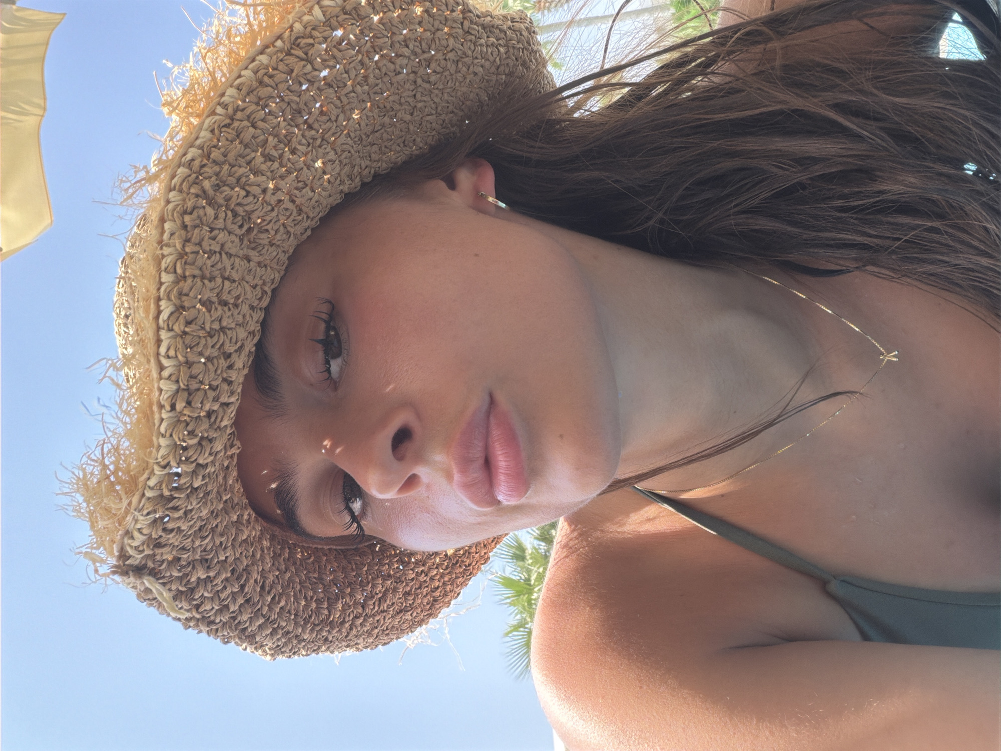 A young woman with dark brown hair, wearing a straw hat and a green bikini top, looks towards the camera with her lips slightly parted. She has a thin gold necklace with a letter "A" pendant. Sunlight creates shadows on her face and hat. She is outdoors, with a bright blue sky above, and palm fronds are visible in the background, suggesting a tropical or Mediterranean location. The scene implies a casual, warm-weather setting.