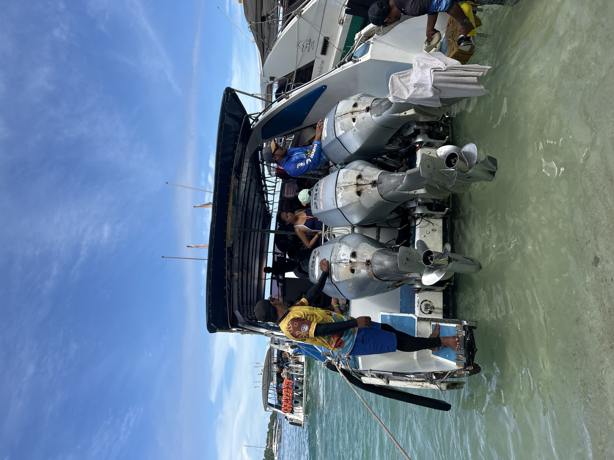 The image depicts the stern of a white speed boat equipped with three large, silver outboard motors in the waters of Ao Nang, Thailand. The motors exhibit wear, and "HONDA 250" is visible on one unit. A man wearing a yellow and brown patterned shirt, blue shorts, and a black cap stands on the port side of the boat's stern platform, hands resting on one of the outboard motors. Another man in a blue long-sleeved shirt and black cap stands to his right, also near an outboard motor. A woman in a white top is visible between these two men. At the far starboard side of the stern, a man in a black shirt and cap bends down, pouring liquid from a yellow container into the turquoise water, beside a white towel draped over a motor cowling. The water is clear, revealing the light-colored sandy bottom near the boat. In the background, under a partly cloudy blue sky, another white tour boat with a stack of orange life vests is visible, with additional boats and sailboats further in the distance.