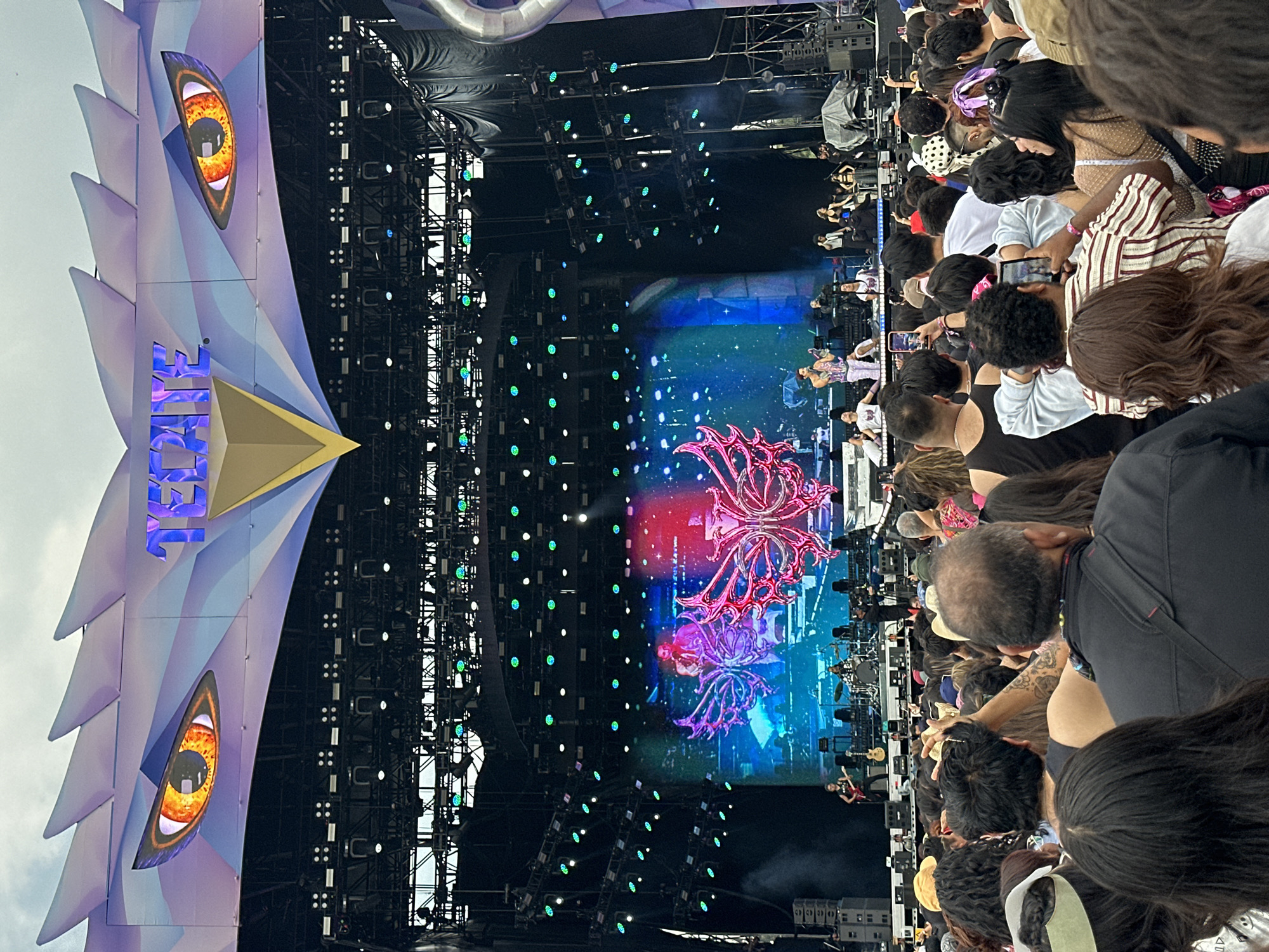 This image captures a concert scene in Mexico City, Mexico, featuring a large crowd watching a performance on a stage. The stage has a complex setup, including lighting rigs and a large video screen displaying graphics. An elaborate structure resembling an eagle's head with illuminated eyes flanks the stage. The crowd is diverse, with individuals of various ages and styles, many of whom are holding up phones, seemingly to capture the event. The scene suggests a large, outdoor music festival or concert event.