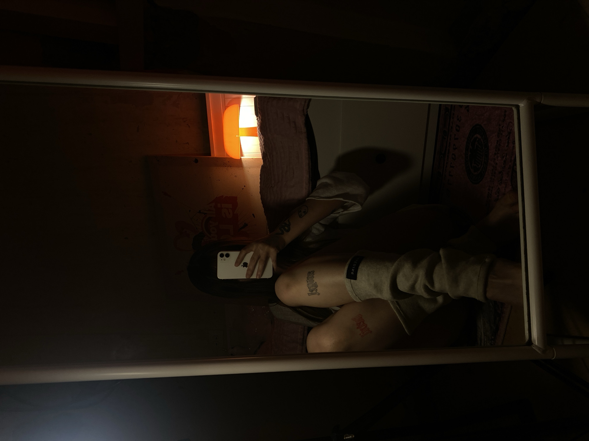 A person is reflected in a white-framed mirror, holding a white iPhone, apparently taking a photo. The person sits cross-legged, visible from the knees down. Tattoos are visible on both legs. One leg has the word "Jopune" tattooed on it, and the other has "Jopa." The room is dimly lit with a small orange lamp visible on the wall. A patterned rug is also partially visible in the reflection. The exact location within Votkinsk, Russia, is indeterminable from the image.