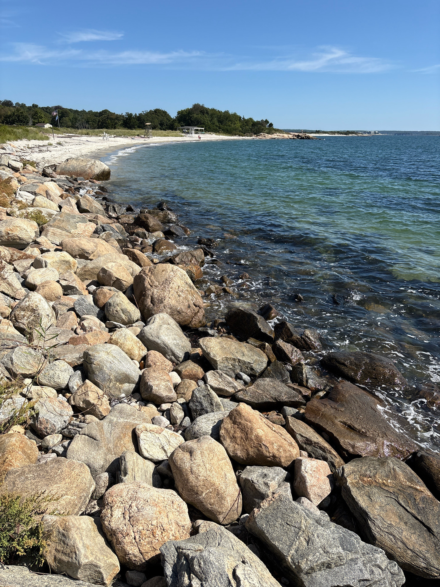 This is a scenic view of a rocky shoreline on a sunny day. In the foreground, a dense collection of various sized and colored rocks fill the lower portion of the image, leading towards the water. The water, a clear turquoise in the shallows and a deeper blue further out, gently laps against the shore, creating small white foam patterns. In the midground, a sandy beach curves into the distance, backed by a line of lush green trees. A few scattered objects are visible on the beach, including two lifeguard chairs and a few people walking along the water's edge. A flagpole stands near the left side of the beach. The sky above is a vibrant, cloudless blue. The location is identified as Waterford, United States of America.