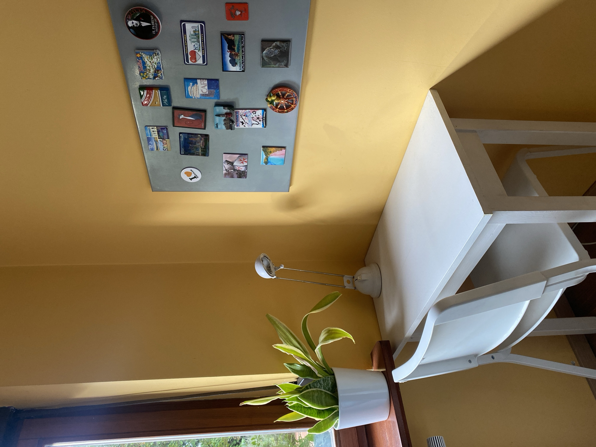 The image shows a corner of a room with yellow walls. A white desk with a white chair is positioned against the wall. On the desk, there is a white plant pot with a snake plant and a white desk lamp. To the right of the desk, a grey magnetic board is attached to the wall, covered with various souvenir magnets from different locations, including "SALOU", "Malta", "Singapore", "PhPhi Island Krabi Thailand", and a magnet with an image of the actor Al Pacino and the text "IL PADRINO SONO IO". There is also a magnet with "I ❤ WROCLAW". A wooden window sill is visible on the left side of the image with a view of greenery outside. The scene appears to be a home office or study area.
