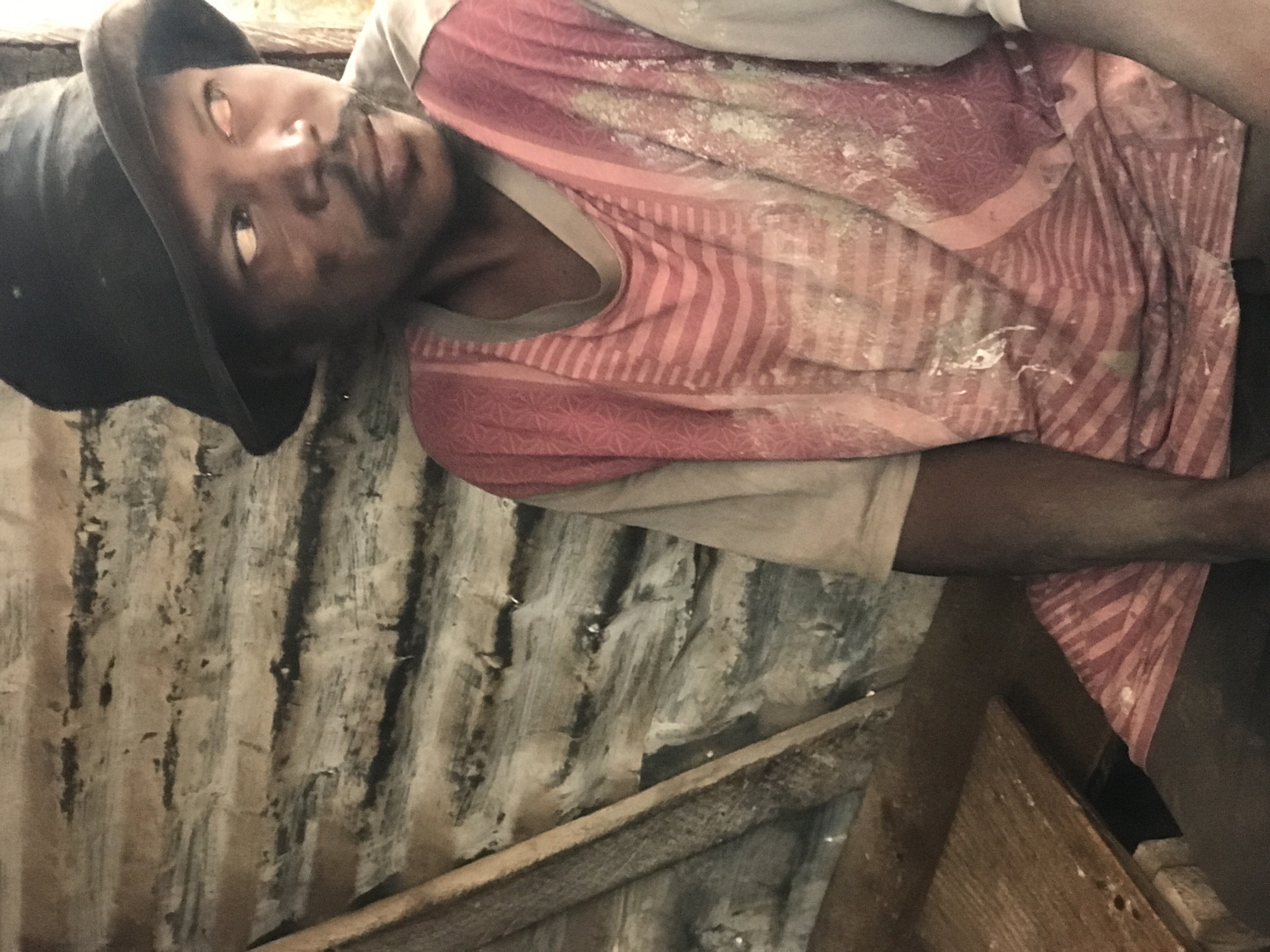 A medium shot captures a man in a black bucket hat and a pink and white striped shirt with a floral pattern on the sleeves, sitting in what appears to be a rustic dwelling in Kabong, Nigeria. The background consists of corrugated metal walls with visible rust and texture, suggesting a weathered environment. A wooden plank runs vertically along the left side of the frame, and a wooden surface is visible in the lower left quadrant. The man's face is partially obscured by the hat, and his gaze is directed to the right. The lighting appears natural, creating shadows and highlighting the textures of the scene. There is a light dusting of what might be powder or dirt on his shirt.