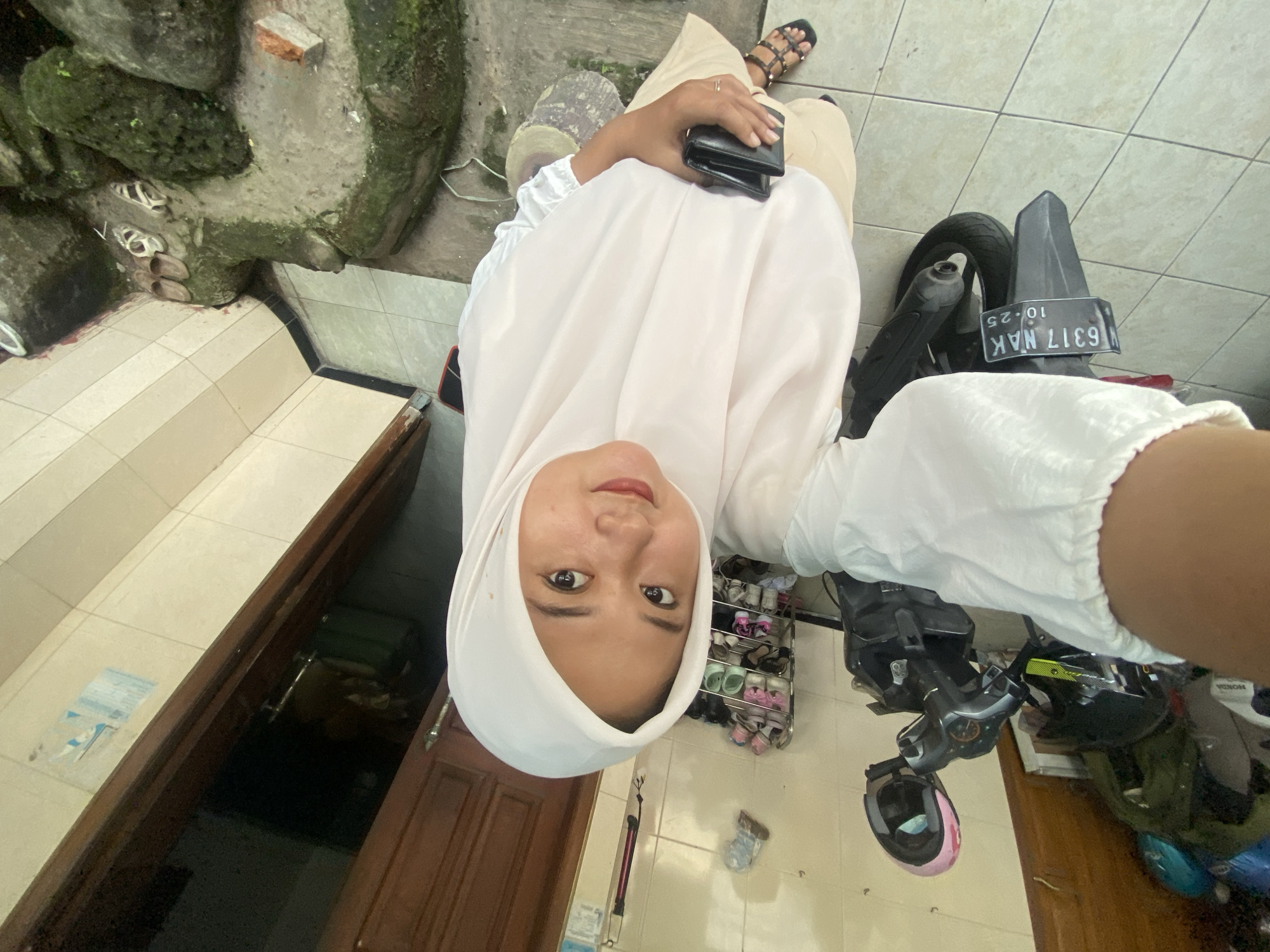 A person wearing a white headscarf (hijab) is taking a selfie. They are standing next to a motorbike with a license plate that reads "G 6317 NAK". The setting appears to be outside a house with a wooden door and a tiled floor. A shoe rack is visible. The location is likely Gelam, Indonesia, based on the license plate prefix "G" which is associated with the Gelam region.