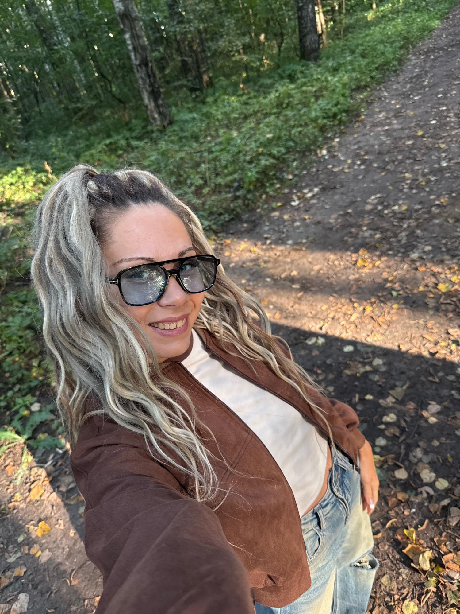 A woman with blonde hair is taking a selfie in a forest. She is wearing sunglasses, a brown jacket, and jeans. The path behind her is covered in leaves and dappled with sunlight. The trees are green and the overall setting suggests an outdoor, natural environment. There is no indication of the specific location within the city of Rusanovo, Russia.