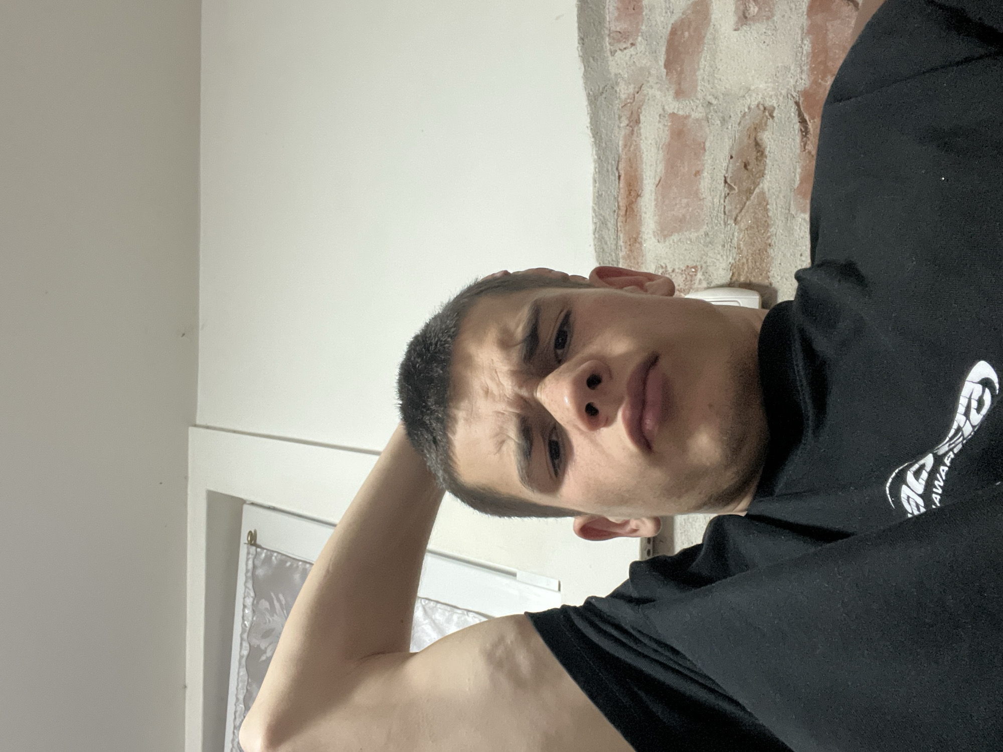 A young adult male is photographed from a low angle, suggesting the photo was taken with a phone held above him. He is wearing a black t-shirt and has a short haircut. His left arm is raised with his hand behind his head. The background includes a white wall and a section of exposed brick. A white window frame with a sheer curtain is visible behind his arm. The exact location within the city of Plovdiv, Bulgaria, cannot be determined from the visual information provided.