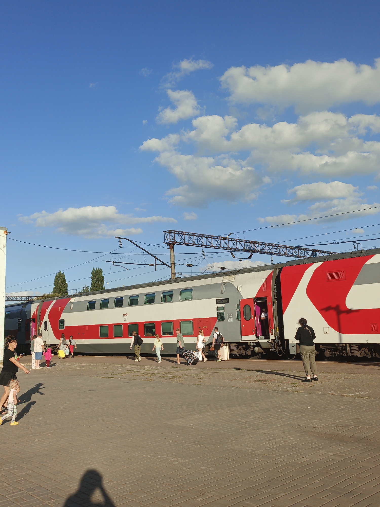 The image shows a double-decker train at a station platform. Several people are visible on the platform, some with luggage, suggesting they are boarding or disembarking. The train is predominantly white with red markings. In the background, there are trees and overhead wires for the train. The sky is blue with scattered clouds. The ground of the platform is paved with bricks. Based on the train livery with the "РЖД" logo, this train is operated by Russian Railways. Given the provided location, this scene is likely taking place at the Gryazi train station in Gryazi, Russia.