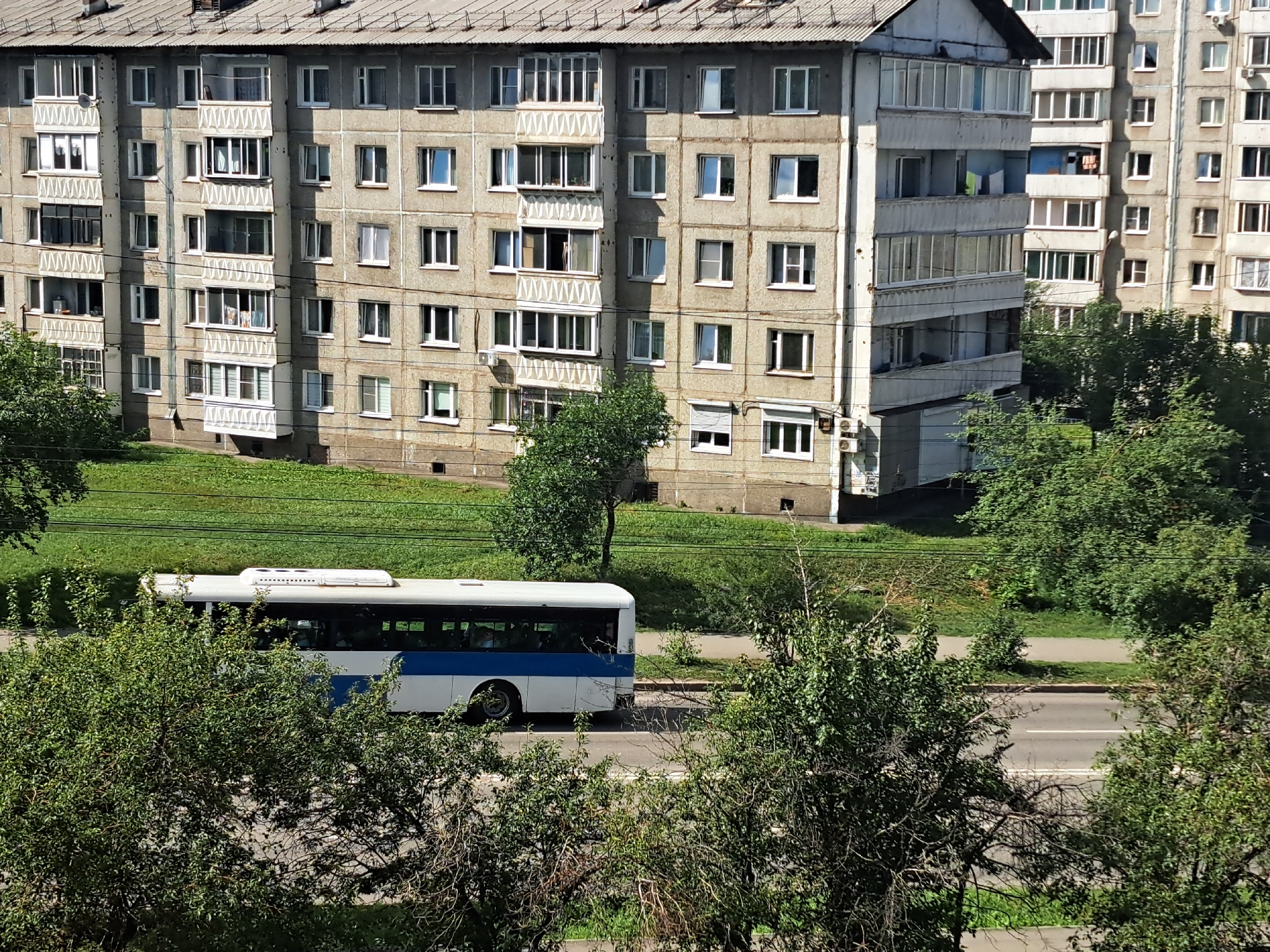 This is a high-angle, full shot of a residential area in Irkutsk, Russia, on a sunny day. The scene is dominated by a large, concrete apartment building with multiple floors and rows of windows, typical of Soviet-era architecture. The building has a grey, weathered facade and a sloping, corrugated roof. Below the building, a green, grassy slope leads down to a paved road where a white bus with a blue stripe is driving. The bus has tinted windows and an air conditioning unit on its roof. Dense green trees and foliage are visible in the foreground and scattered throughout the scene, partially obscuring the view of the road and the lower levels of the apartment building. The sky is not visible.