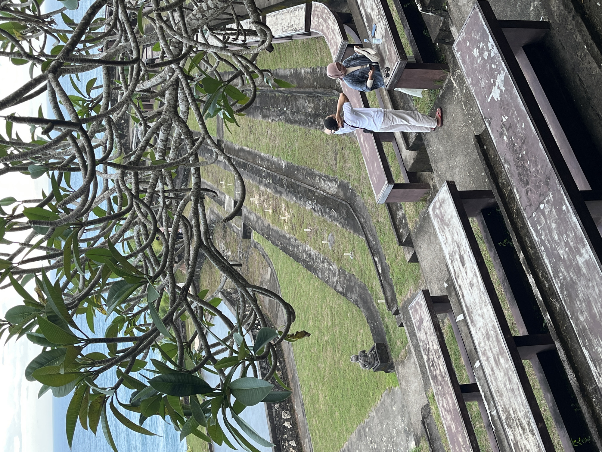 The image shows an elevated view within the Garuda Wisnu Kencana Cultural Park in Kuta Selatan, Indonesia. Stone benches line the right side of the frame, leading to an open grassy area divided by concrete pathways. Two people occupy a bench in the middle distance; one in white clothing stretching, the other with a head covering. A tree with prominent branches and green leaves dominates the left foreground. The sea is visible in the distance.