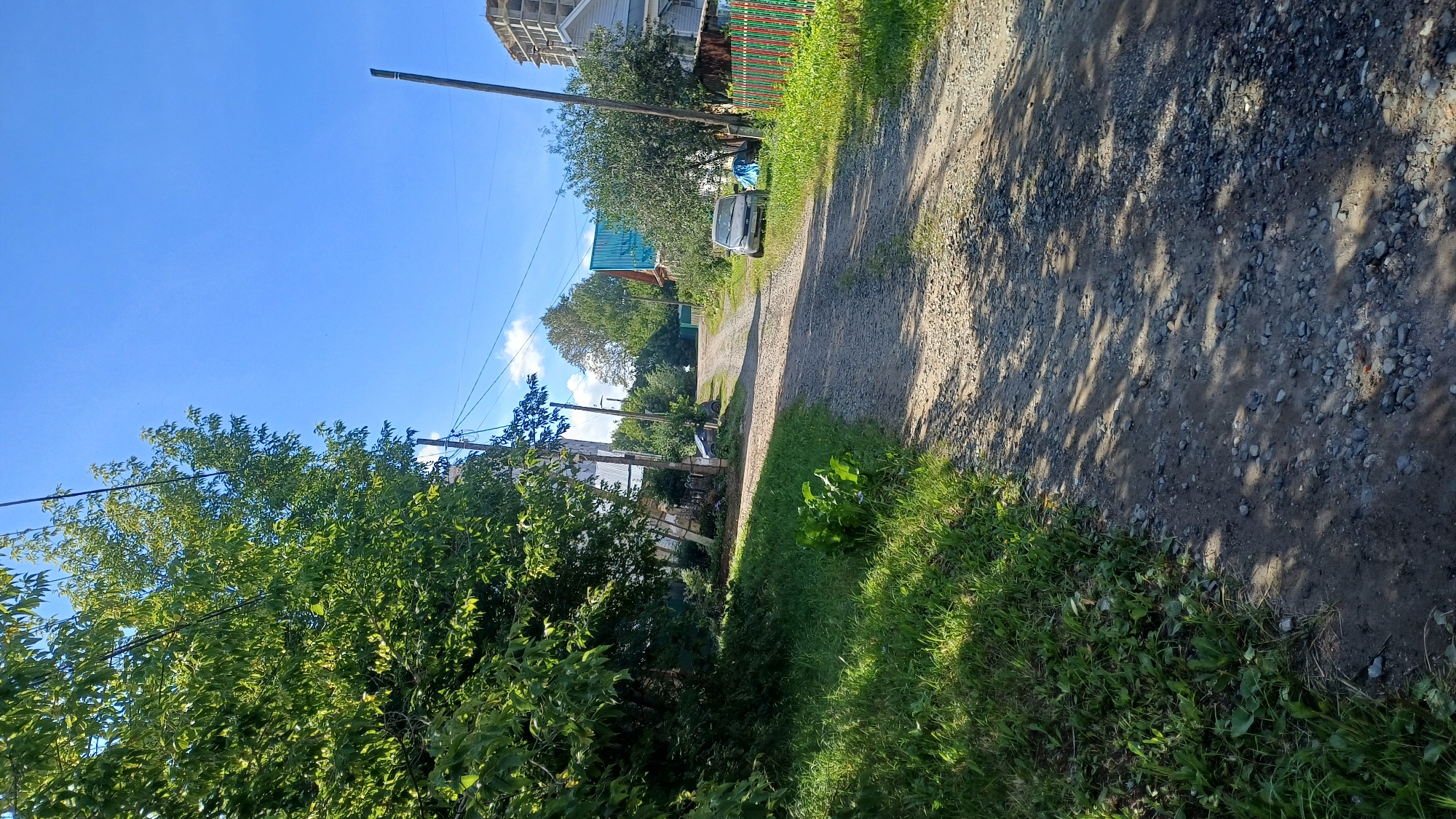 The image depicts a rural road with a gravel surface, flanked by lush green vegetation on the left and houses on the right. The road curves gently into the distance. In the foreground, shadows of tree branches are cast across the road and the grassy verge. A large tree with dense green foliage dominates the left side of the frame. On the right side of the road, there are several houses, one with a distinctive teal-colored roof. A multi-story building under construction is visible in the background. Two cars are parked along the right side of the road, one in the middle ground and another further back. A colorful fence with green and red vertical slats runs along the edge of a property on the right. The sky is a clear, bright blue with a few wispy clouds. The scene appears to be a sunny day in a village or suburban area. This location is likely a residential street in Izhevsk, Russia, characterized by its blend of natural elements and modest housing.