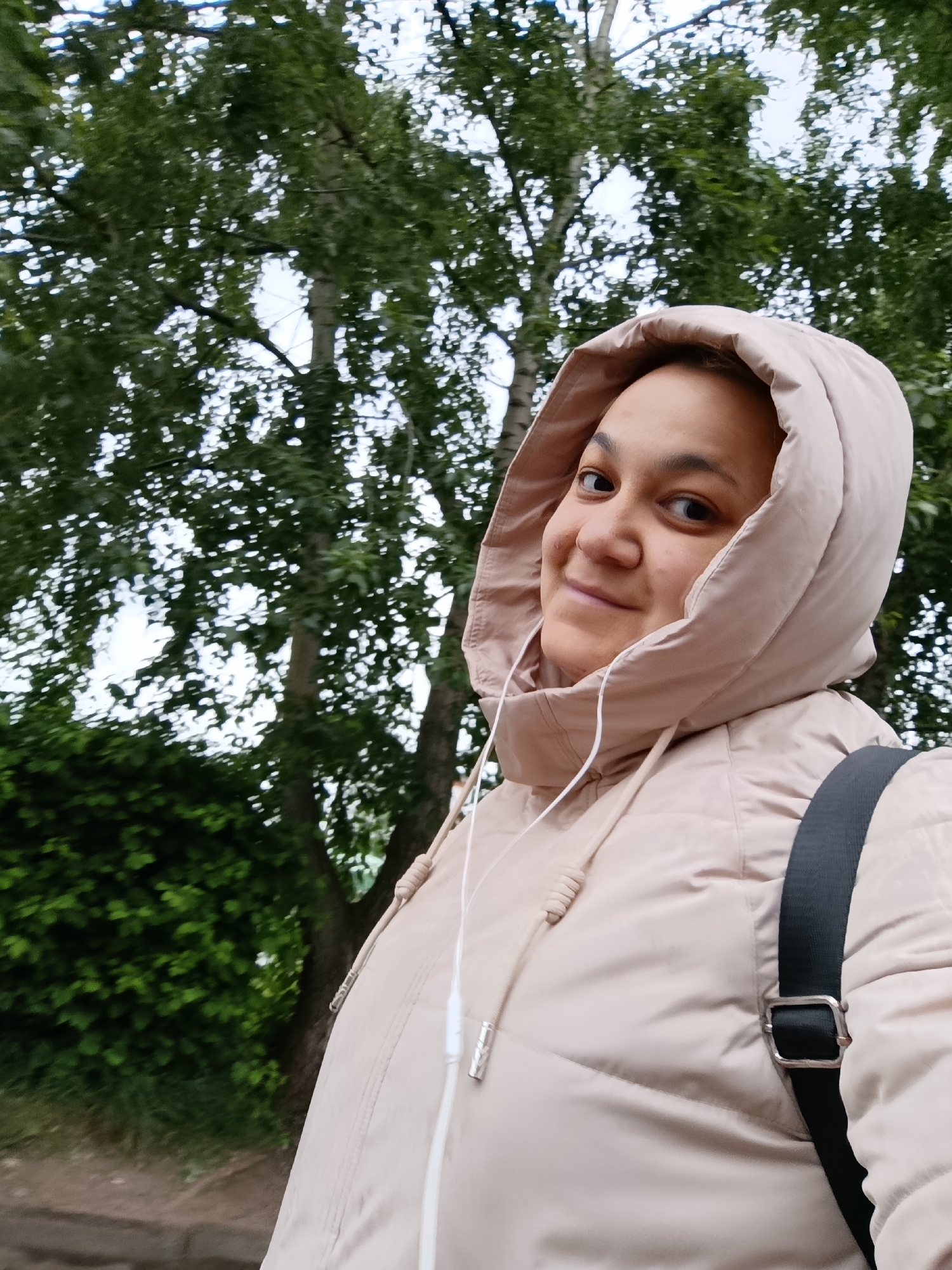 A close-up outdoor portrait features a young person looking towards the camera with a gentle smile. They are wearing a light beige, possibly padded, hooded jacket with the hood up, framing their face. White wired earphones are visible around their neck and leading into their ears, suggesting they are listening to music or a podcast. A black strap from a backpack is visible over their right shoulder. The person's expression is friendly and content.
The background is dominated by lush green foliage and tree trunks, indicative of a natural outdoor setting, likely a park or a treelined street. The leaves appear to be on deciduous trees, possibly birch given the light-colored trunk visible in the upper background. The sky, visible through gaps in the leaves, is overcast and bright, suggesting daylight. The lighting is soft and diffused. In the bottom left, a dark patch of ground, possibly a wet path or dirt, is visible. There is no visible text in the image. The scene conveys a calm, casual moment outdoors, possibly in Chaikovsky, Russia.