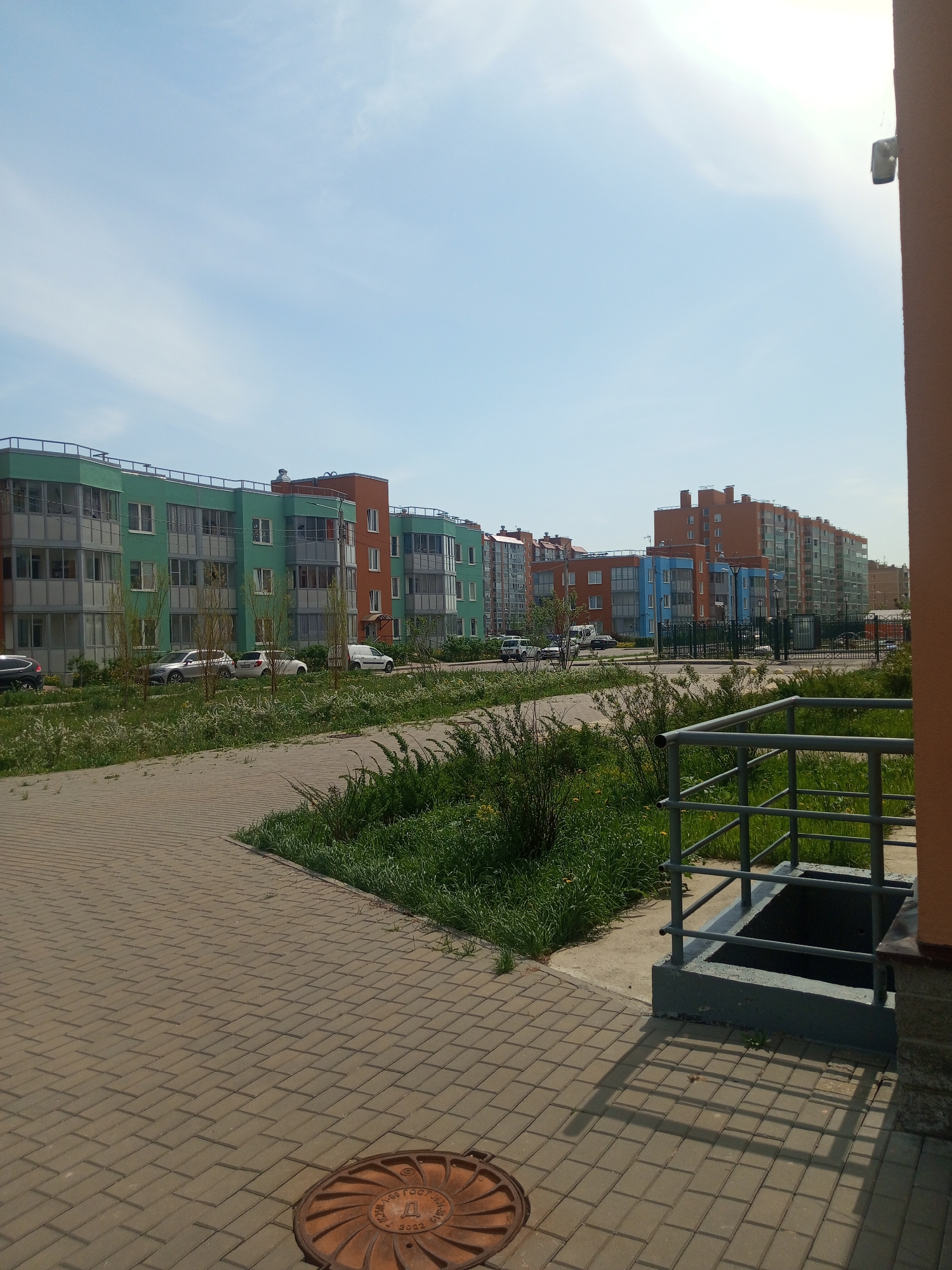 The image depicts a street scene in Kommunar, Russia, showcasing a row of colorful, modern apartment buildings. The buildings are painted in shades of green, orange, and blue, with multiple stories and visible balconies. Several parked cars are visible in front of the buildings.
A paved brick walkway is in the foreground, leading into the scene. To the right, a set of metal railings surrounds a sunken area, likely for utility access. Patches of green grass and foliage are scattered throughout the scene. A manhole cover is visible on the left side of the walkway. The sky is clear and blue, suggesting a sunny day. The overall atmosphere is calm and residential.