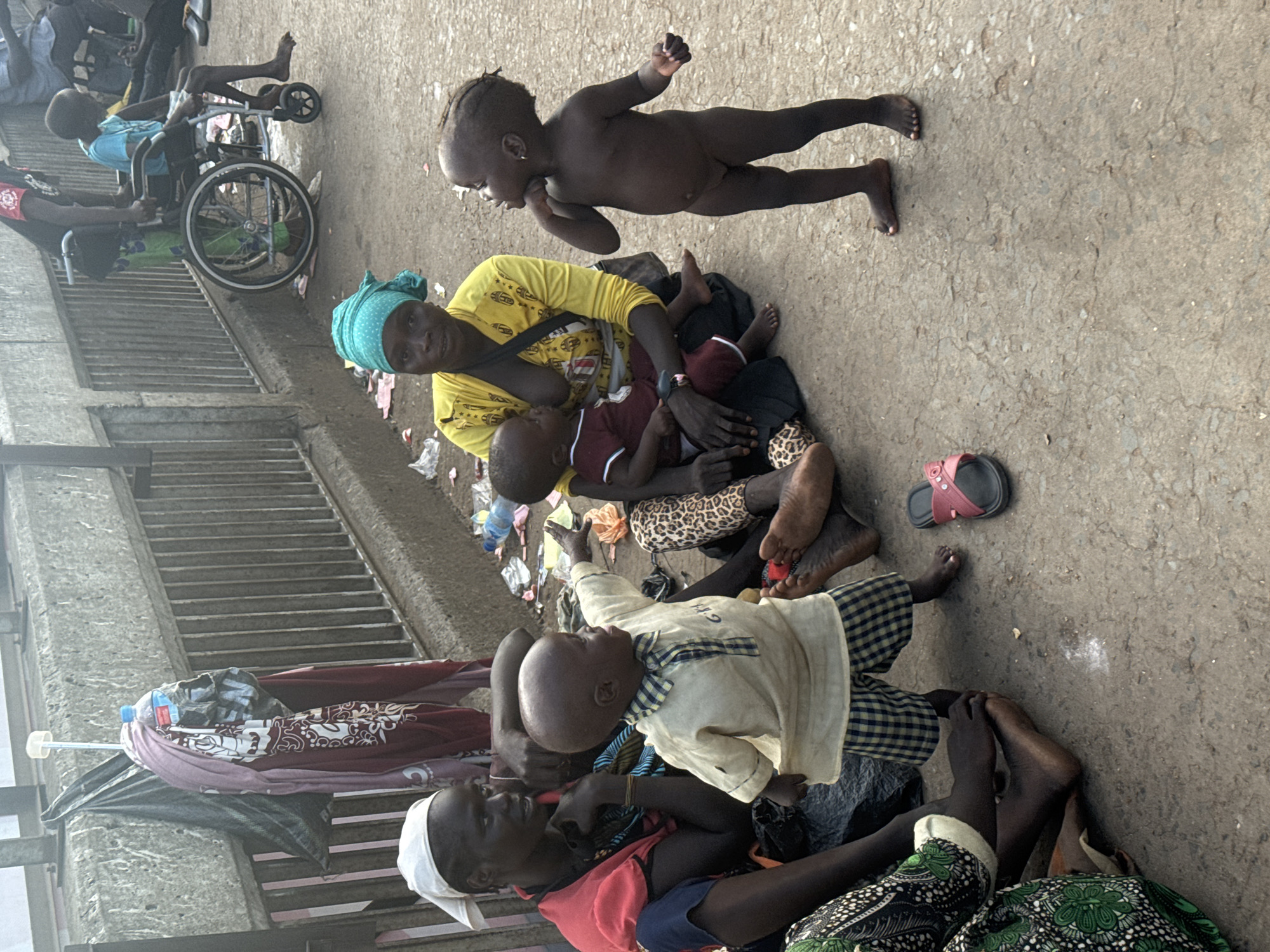 The image depicts a scene on a busy street in Onitsha, Nigeria, where several individuals, including women and children, are gathered. A woman wearing a yellow shirt and a blue headscarf is seated, holding a baby. Another woman in a red shirt and blue trousers is also seated, with a child standing in front of her. A young, unclothed child stands to the right, gesturing with an open hand. In the background, a person in a wheelchair is being attended to by another individual. The ground is paved with concrete and littered with various items, including plastic bottles, papers, and a pair of pink sandals. A metal railing runs along the side of the walkway, behind which is a concrete barrier. The overall atmosphere suggests a bustling urban environment, likely a public space where people are seeking shelter or engaging in daily activities.