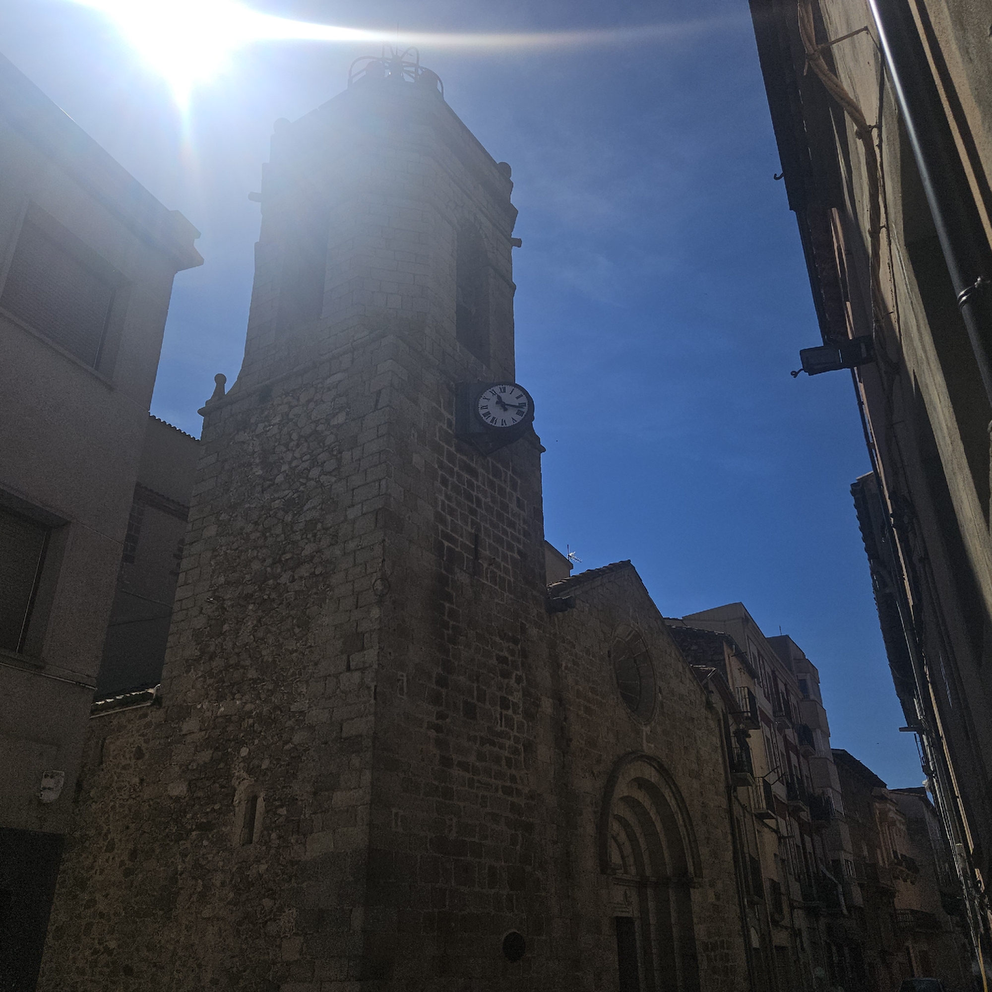 A ground-level view of the city of La Jonquera, Spain, in daylight showcases a building made of tan stone and mortar construction. The building has a prominent tower affixed with a clock. The clock is high up on the building's surface and has a white clockface with Roman numerals and black hands. The tower is square and stacked with blocks of stone. A clear sky can be seen around the tower. The building's architectural style is Gothic, with a rounded arch over the entrance. A street flanks each side of the building, with other buildings parallel to the pictured one. The sun is above the scene, with the building primarily in shadow.