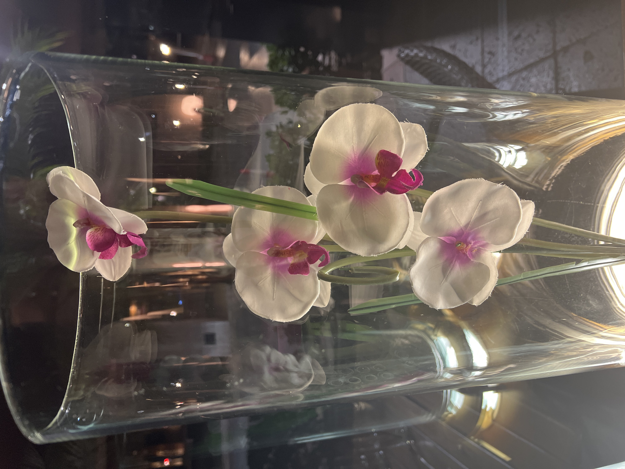 The image is a close-up shot of artificial white orchids with fuchsia centers displayed in a clear glass cylinder vase. The vase is tall and cylindrical, with visible reflections of lights and possibly foliage in the background. Four orchid blooms are visible, with stems and green artificial leaves. The lighting suggests an indoor setting, likely at night, with some bokeh effects in the background. The location is not specified in the image.