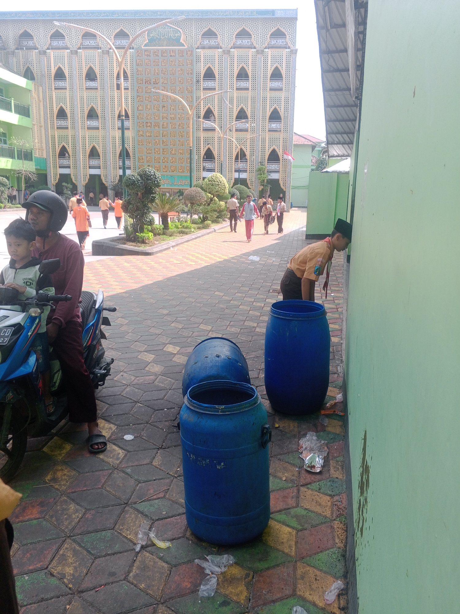 The image depicts an outdoor scene in what appears to be a schoolyard or courtyard in Ponorogo, Indonesia. In the background, a large, ornate building with a yellowish facade and intricate architectural details is visible. The building has multiple arched windows and decorative patterns. In the foreground, several people are present. On the left, a man wearing a dark helmet and a maroon shirt is seated on a blue motorcycle, with a young boy in a light-colored shirt sitting in front of him. To the right, a young person in a scout uniform, consisting of an orange shirt and brown pants, is leaning their head against a light green wall. In front of this individual are two large blue plastic barrels. Scattered on the patterned pavement around the barrels are pieces of plastic trash. Other individuals, possibly students, are seen walking in the middle ground of the image, some wearing uniforms that include orange or white tops and dark pants. The pavement is made of interlocking geometric tiles in various colors, including yellow, red, green, and gray. Trees and manicured bushes are visible in the mid-ground between the people and the large building. The overall lighting suggests it is daytime, likely sunny.