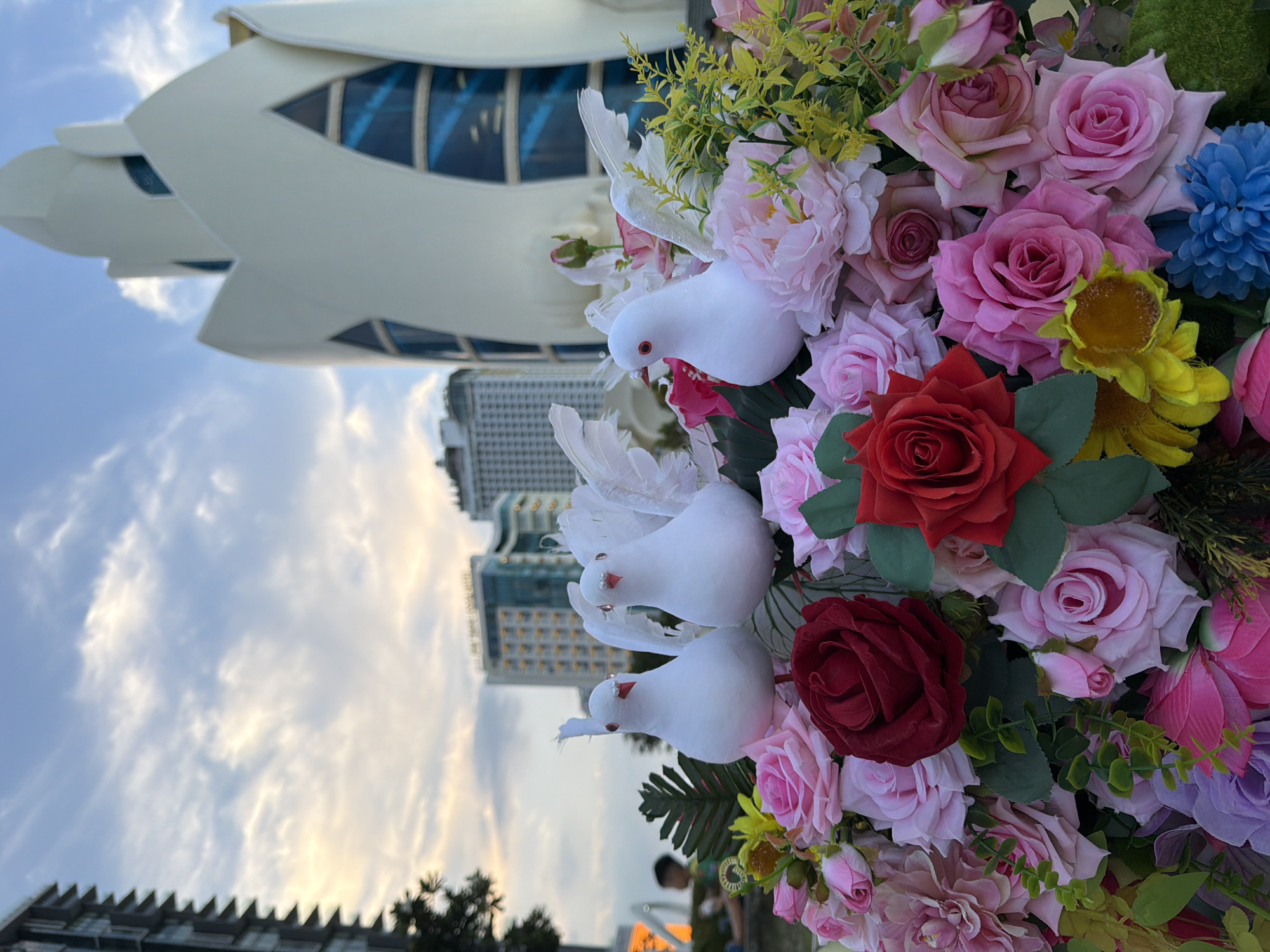 A close-up, eye-level shot showcases a vibrant arrangement of artificial flowers and three white doves with red beaks and eyes, positioned in the foreground. The flowers are a mix of roses in various shades of pink, red, and purple, with some yellow sunflowers and a blue pom-pom-like flower. Behind the floral arrangement, a striking, petal-shaped white building with large blue-tinted windows dominates the midground. In the background, several modern high-rise buildings and a cloudy sky are visible. A blurry figure of a child can be seen sitting on the ground to the left, partially obscured by a leafy plant. The scene appears to be an outdoor public space, possibly a park or plaza, in Nha Trang Ward, Vietnam, given the architectural style of the prominent building and the urban setting.