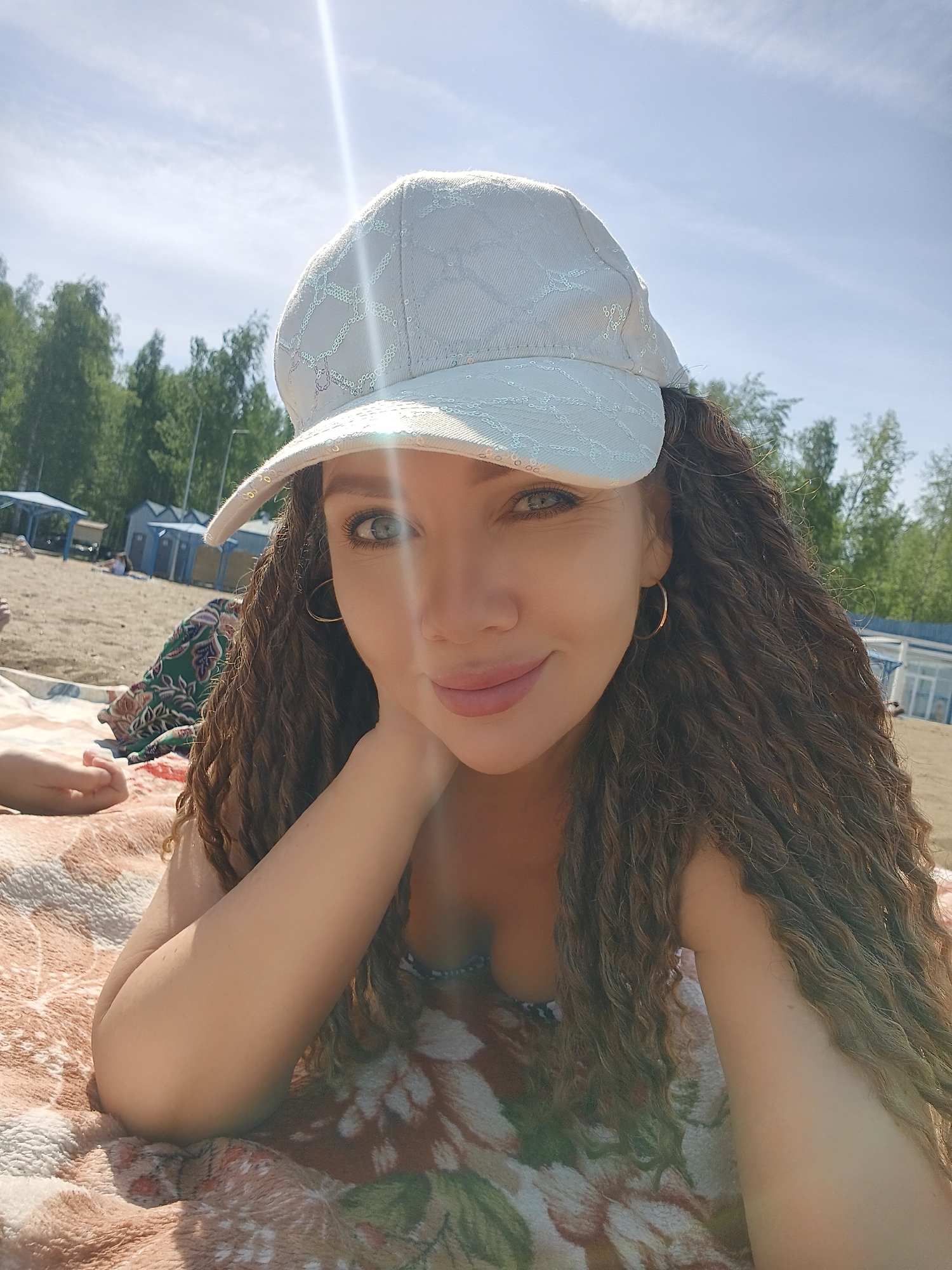 A woman with curly brown hair and a light-colored cap is lying on a patterned blanket on a beach. She is resting her head on her hand and looking at the camera. In the background, there is sand, trees, people, and some beach structures. The location is a beach, likely near a body of water, in a city that cannot be determined from the image.