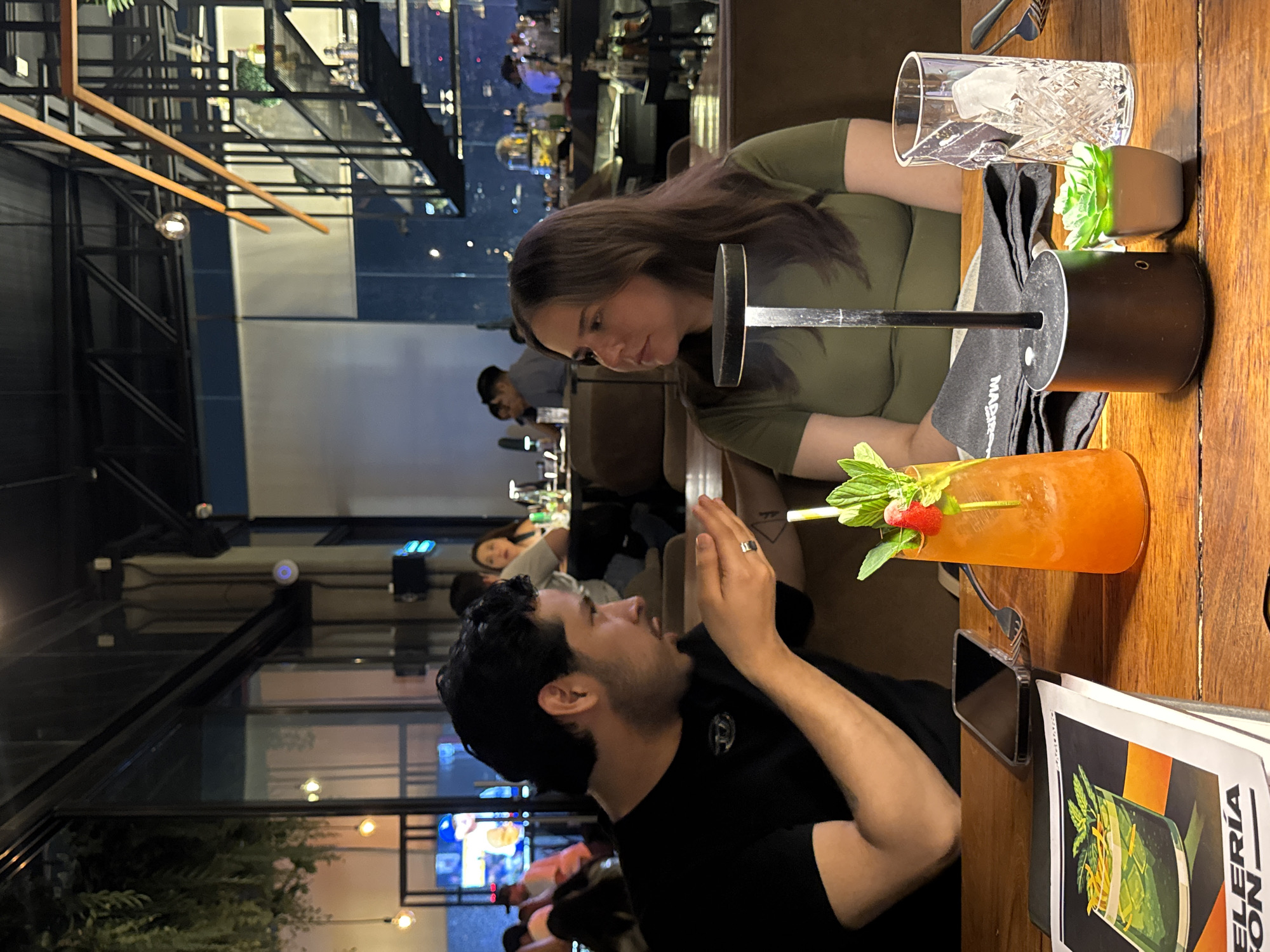 A candid, slightly rotated, medium shot shows a couple at a restaurant in Puebla, Mexico. A dark-haired man in a black t-shirt faces a woman wearing an olive green shirt. They sit closely together. The man is gesticulating with his right hand.
On the wooden table in front of them are a clear cocktail glass with ice, a small planter with greenery, a cocktail with fruit garnishes, and a menu titled "Cocteleria Con". A cell phone is placed near the menu and cocktail.
The restaurant has an open, modern design with exposed metal beams and glass walls. The background features other patrons at tables and views of the city at night. Through the glass wall, a television screen is visible.