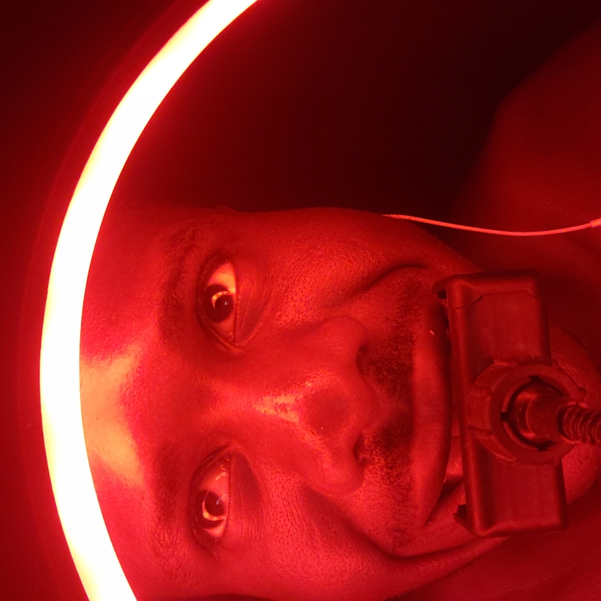 The image captures a close-up, upward-angled view of a person's face, predominantly illuminated by a striking red light. The subject appears to be an individual with dark skin, exhibiting some facial stubble or a short beard. Their eyes are open and look directly towards the camera, maintaining a neutral expression.
A prominent, bright, curved light source, appearing yellow-white, forms an arc along the left side of the frame, casting a strong glow on the left side of the person's face. Near the person's chin and mouth area, a dark, rectangular device is visible, connected by a cable or tube. Another thin, white wire or tube can be seen extending from the upper right portion of the image.
The scene is dominated by the intense red lighting, which creates a somewhat clinical or specialized environment. The background is dark and indistinct, suggesting an enclosed indoor space. The person seems to be undergoing a procedure or treatment, possibly involving light therapy or a medical/dental apparatus. No specific time of day or external location cues are visible within the image, though the provided context places it in Narayi, Nigeria. There is no visible text in the media file.