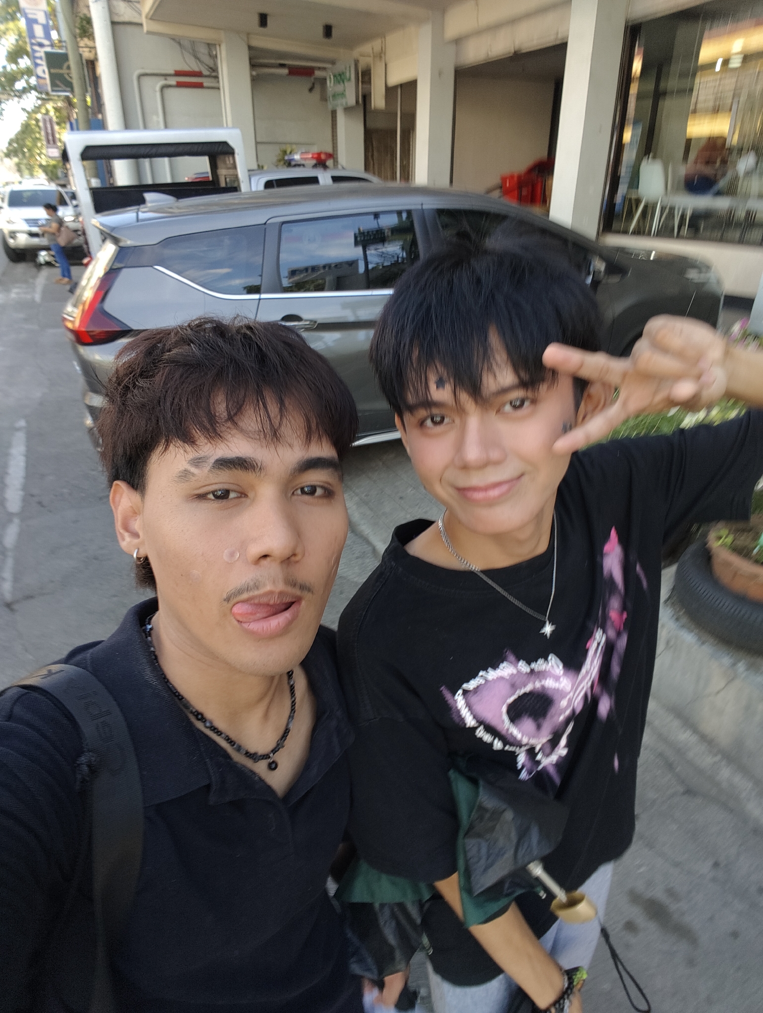 In an outdoor urban setting, likely within Davao City, Philippines, two young Asian individuals are captured in a selfie. The person on the left, with dark hair, is positioned closer to the camera, sticking out their tongue with a playful expression. They are wearing a black collared shirt and a beaded necklace. The person on the right, also with dark hair, is smiling and making a peace sign with their fingers. They are wearing a black t-shirt with a graphic design and a silver necklace. Behind them, a dark-colored van is visible, suggesting a street or parking area. In the background, parts of a building and other vehicles are present. The overall scene appears to be a casual moment captured on a sunny day.