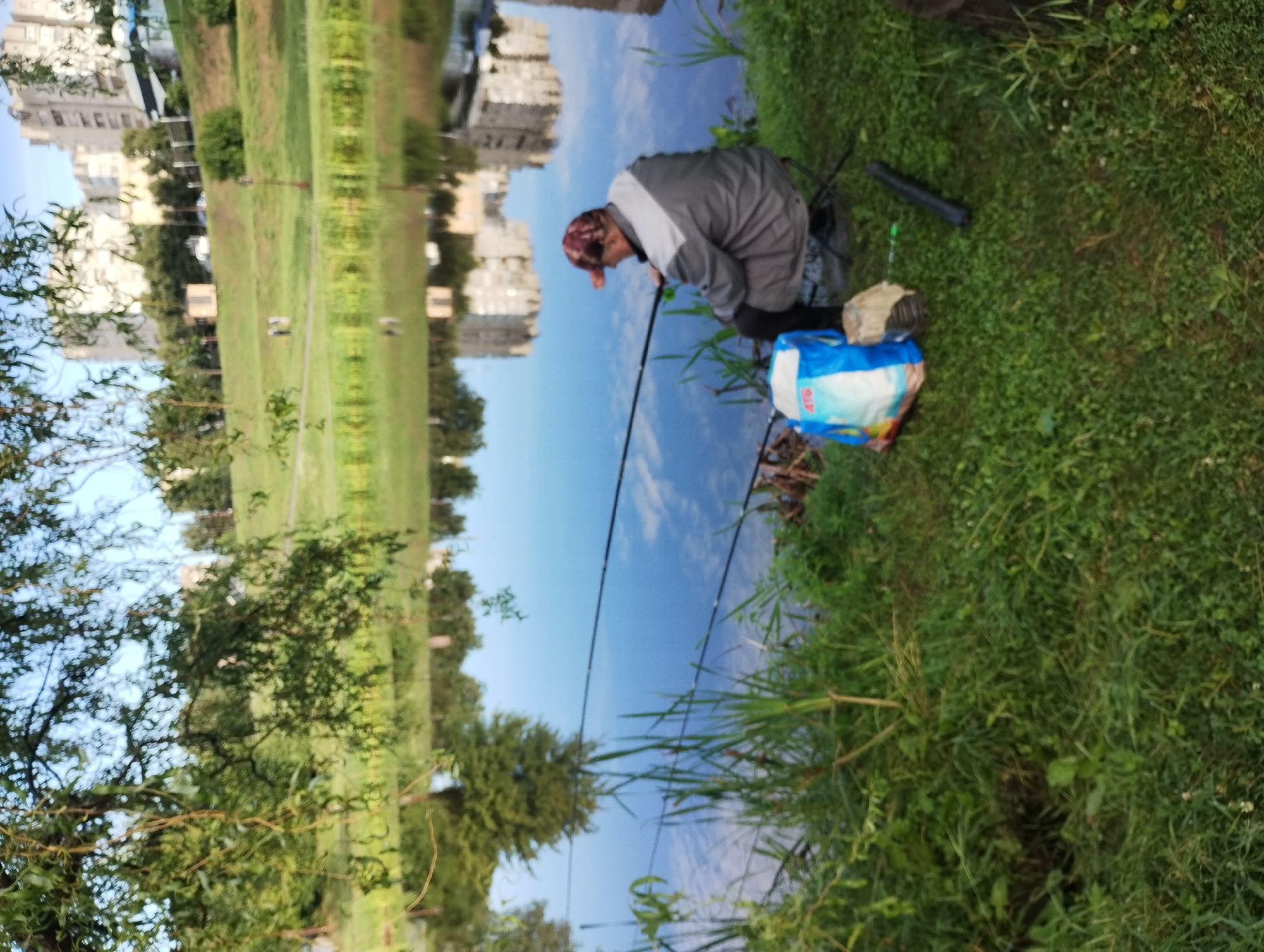 An elderly man is fishing on the bank of a calm river, possibly the Psel River in Sumy, Ukraine. The man is seated on a small foldable stool, facing away from the camera, wearing a patterned cap and a grey and white jacket. His fishing rod is extended over the water, and a blue and white bag lies at his feet. The water reflects buildings and trees from the opposite bank, suggesting an urban or suburban setting. The scene is captured in daylight, with lush green grass and reeds along the water's edge.