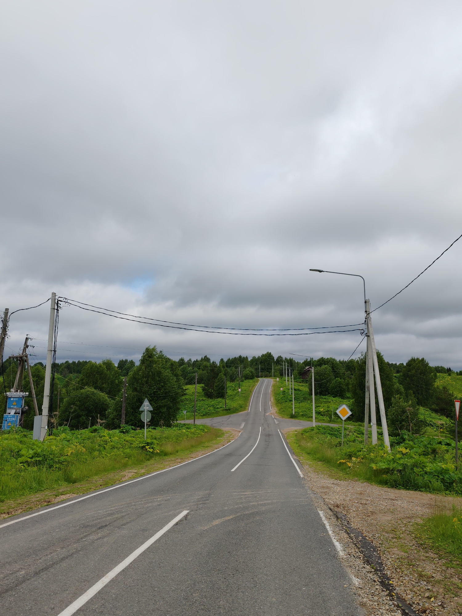 The image shows a two-lane asphalt road in the village of Chukhlem, Russia. The road curves and ascends a hill. On either side of the road, there is lush green vegetation, including trees and tall plants. Several utility poles with electrical wires run alongside the road. There are traffic signs present, including a diamond-shaped sign with a yellow background and a black border, and a triangular sign with a red border and a white background. The sky is overcast with gray clouds. There are no people or vehicles visible in the image. The scene appears to be a rural landscape on a cloudy day.