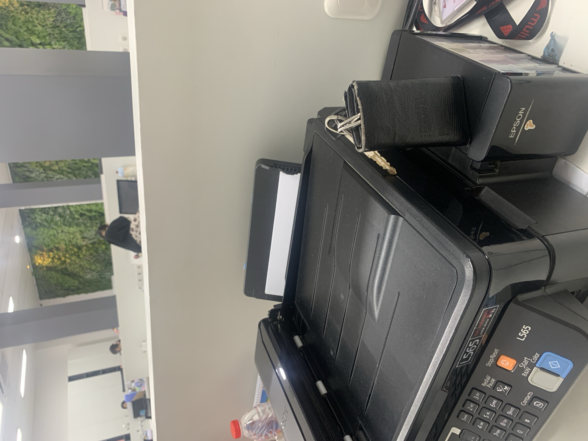 The image shows a close-up of a black Epson L565 printer with its scanner lid open. A black leather wallet with keys attached is resting on top of the printer. To the left of the printer, a red-capped plastic water bottle is visible. In the background, there is a glimpse of an office environment with people and a green wall feature. The scene is indoors, likely in an office setting. There is no specific indication of the location being in Palembang, Indonesia.