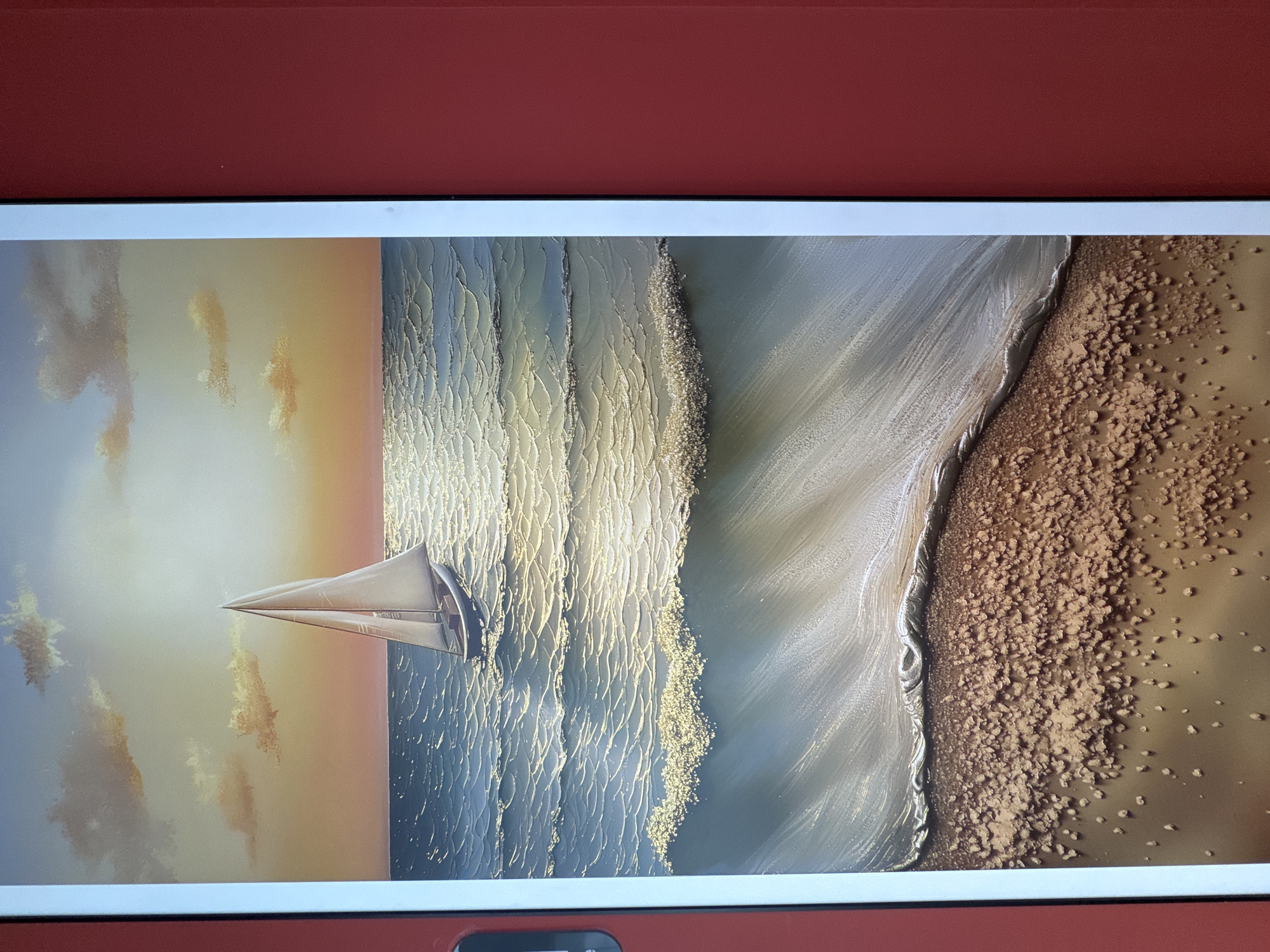A painting of a sailboat on the ocean at sunset is displayed. The painting is framed by a white border and a red wall. The sailboat is white with a single sail. The ocean is a deep blue with cresting waves. The sunset is a warm orange and yellow, casting a golden light on the water. The beach in the foreground is sandy with a textured appearance.