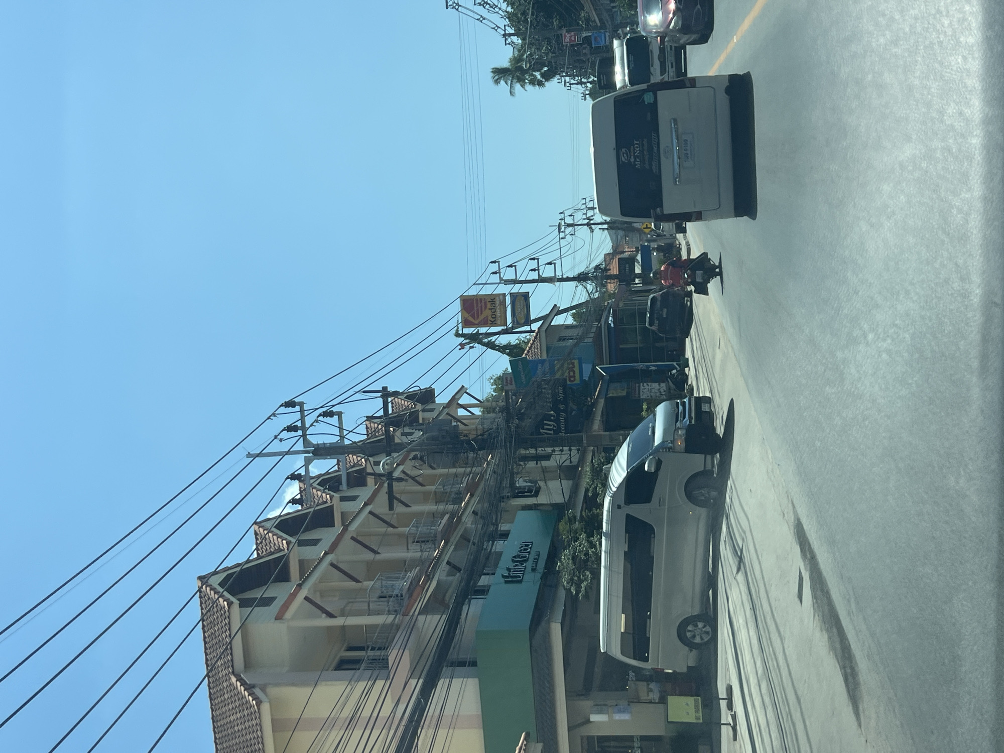 This is a photograph taken from inside a vehicle, looking forward and slightly to the left at a street scene in Ko Samui District, Thailand. The sky is clear and bright blue. On the left side of the street, a multi-story building with a tiled roof and balconies is visible, with numerous overhead power lines and electrical poles in front of it. Signs for "Little Green" and "My. Spa Beauty & Spa" are affixed to the building. A white minivan is parked on the left side of the road, and a dark-colored sedan is in front of it.
The road itself is paved and appears to be two lanes in the direction of travel. A white minivan is in the right lane, further down the road. A person on a red motorcycle is ahead of the minivan in the left lane. Other vehicles, including a white truck and a red car, are visible further down the road. Palm trees and other vegetation are present along the right side of the street. The overall impression is of a busy commercial street under a bright, sunny sky.