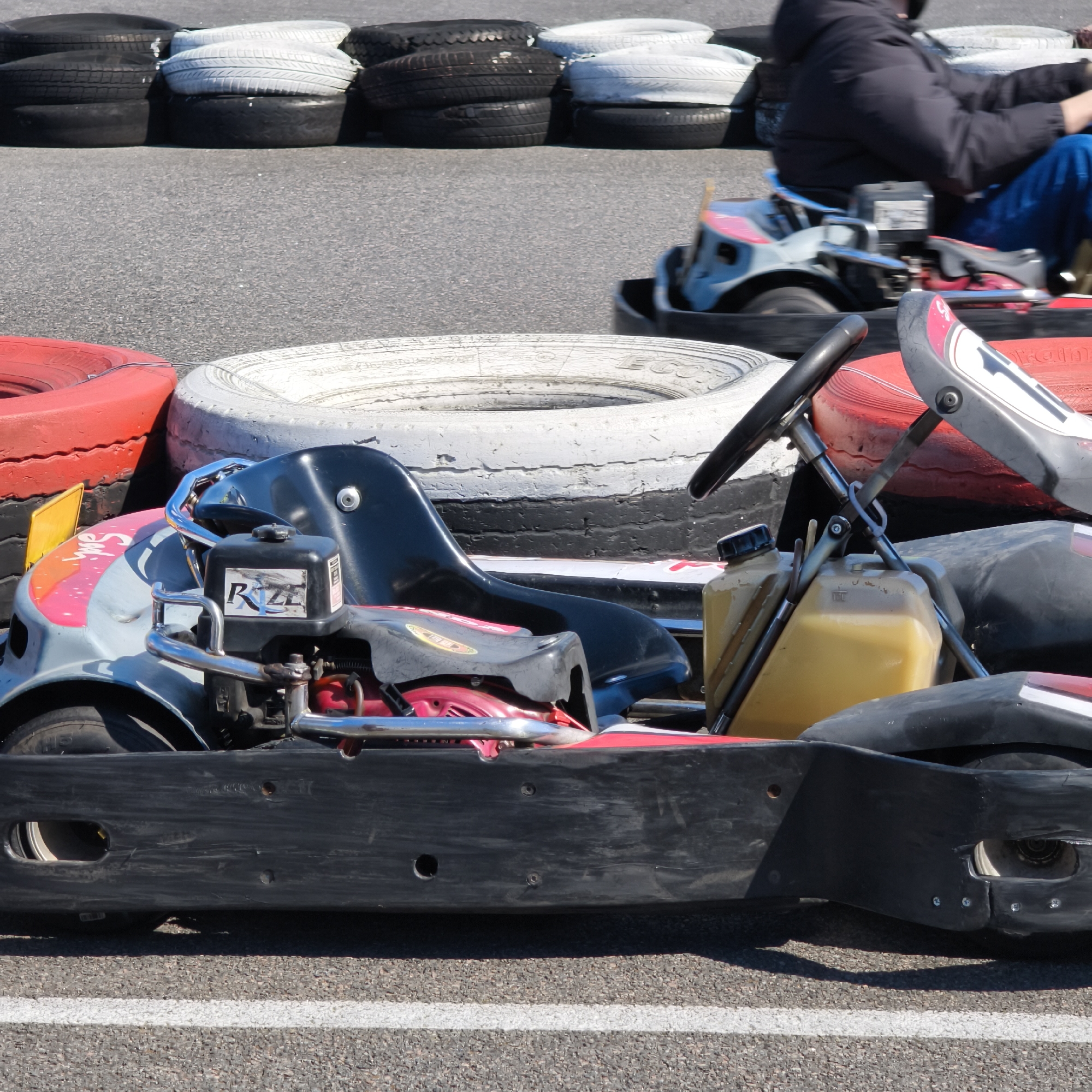 The image shows a go-kart track, likely in Hrodna, Belarus, as specified. A stationary go-kart is in the foreground, positioned near a barrier of stacked tires painted in white, red, and black. A second go-kart, with a driver in a dark jacket and blue pants, is moving on the track in the background. The track is asphalt with a white line marking a boundary. The foreground go-kart has a black seat, a yellow fuel tank, a black steering wheel, and an engine labeled "Rize."