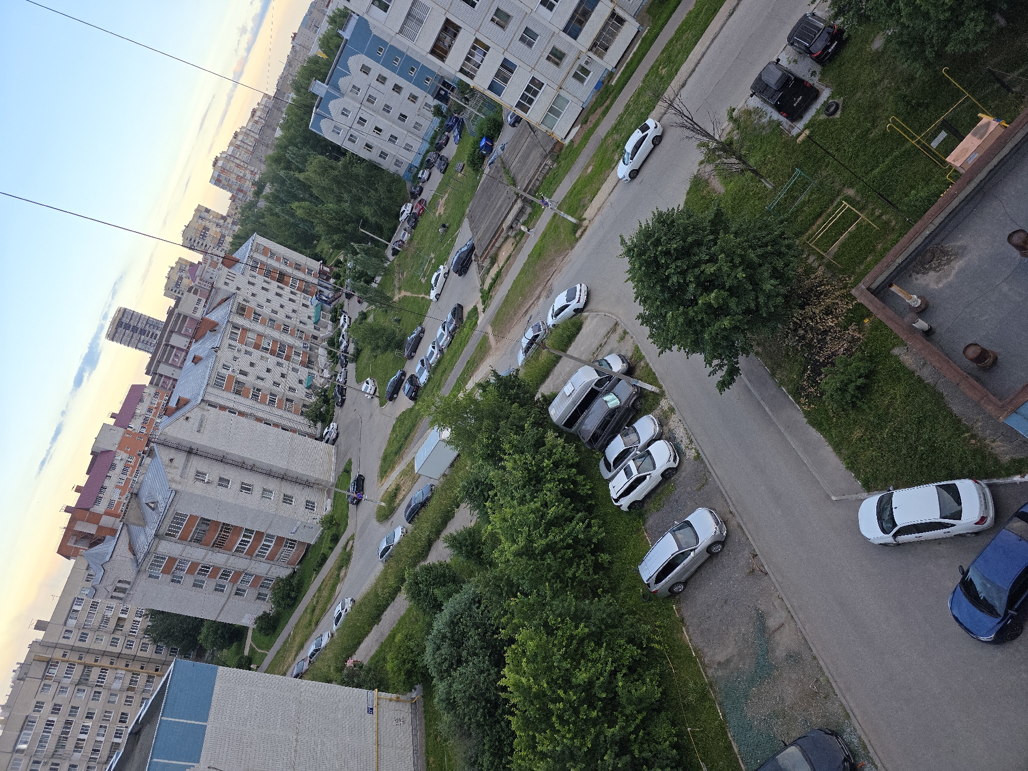 The image depicts a residential area in Novocheboksarsk, Russia, captured from an elevated perspective, likely a balcony or window. The scene is dominated by multi-story apartment buildings with light-colored facades and balconies. Several cars are parked on the asphalt roads and in designated parking areas between the buildings. Green trees and grass are interspersed throughout the urban landscape, indicating a well-established residential development. The sky is visible in the upper portion of the image, with a hint of sunset colors. There are no visible people or animals in the frame, and the overall atmosphere suggests a quiet, everyday urban scene.