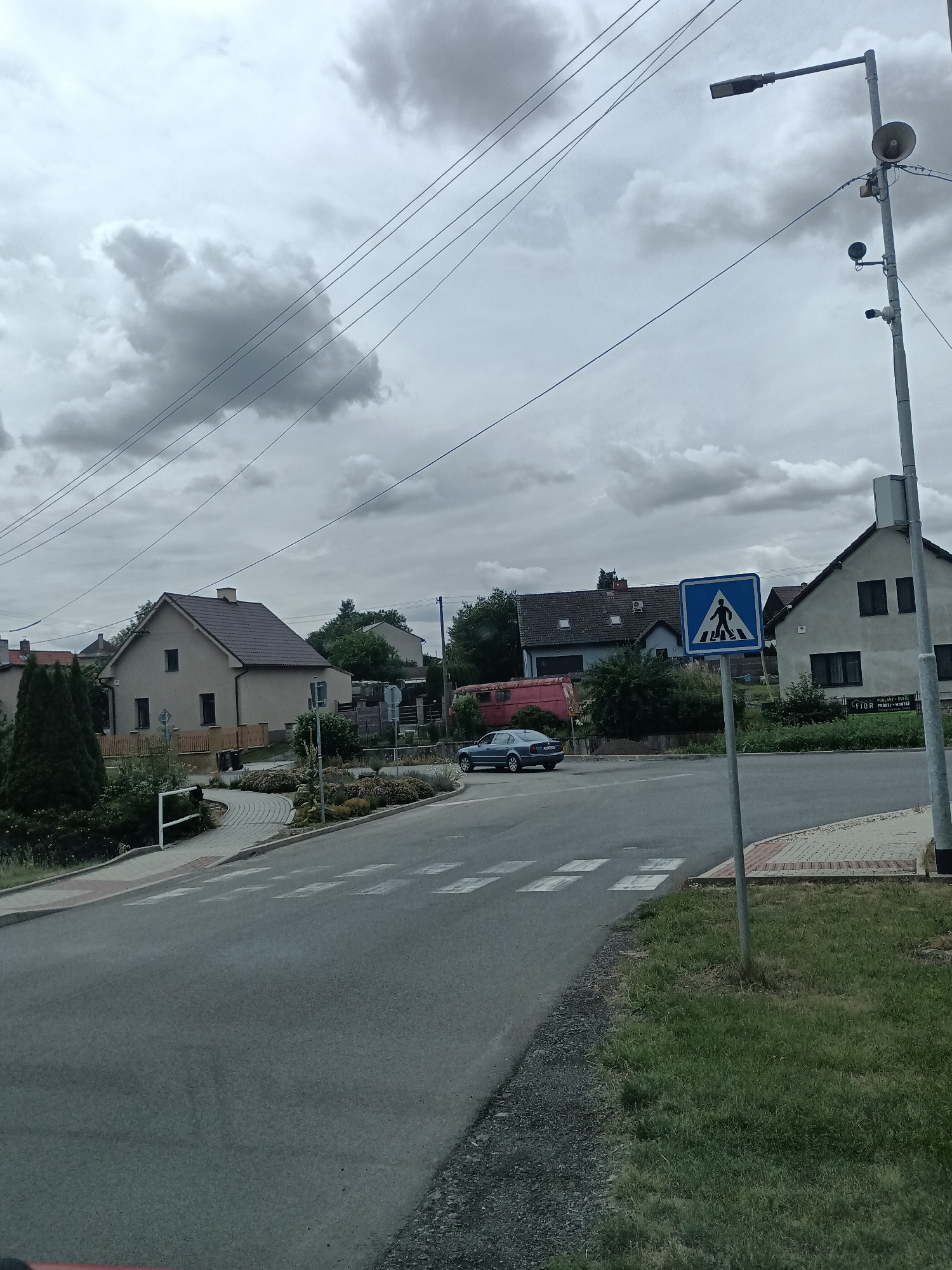 The image depicts a cloudy day scene in the city of Čistá, Czechia, focusing on a street intersection with a pedestrian crossing. A dark blue sedan is seen driving across the pedestrian crossing. To the right of the crossing, a blue and white pedestrian crossing sign is mounted on a pole. Behind the sign, a building with dark windows is visible. A street lamp with multiple attached devices, including what appears to be a speaker and cameras, is also present on the right side of the image. The road surface is asphalt, with white markings for the pedestrian crossing. Beyond the road, there are houses, trees, and a paved walkway. The sky is overcast with dramatic, dark clouds.