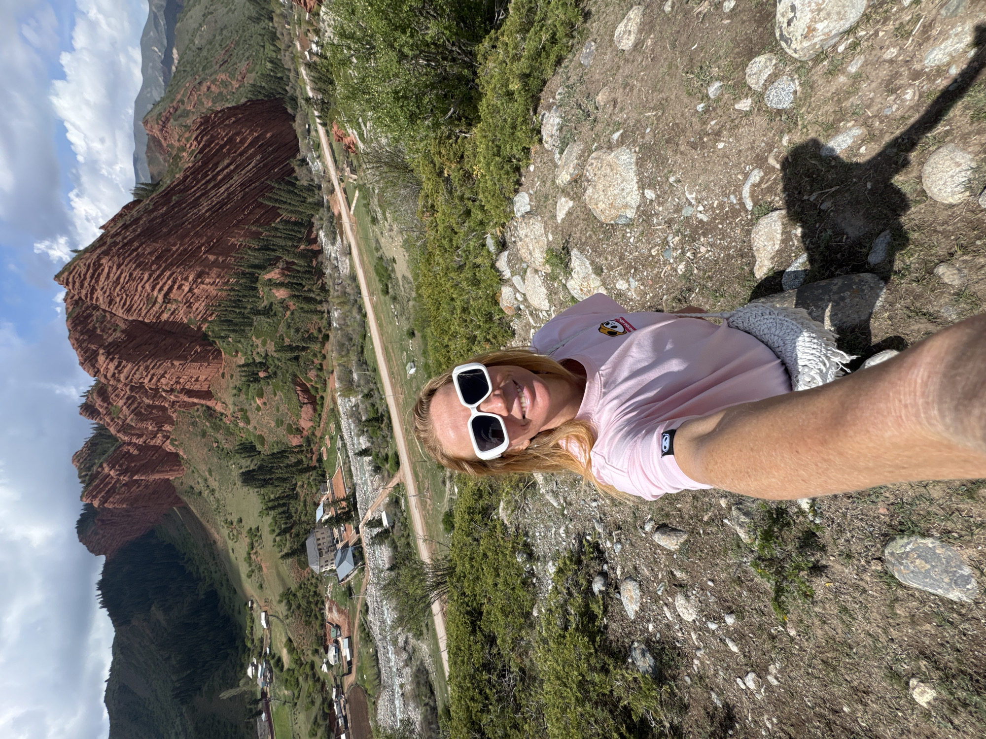 An outdoor selfie captured from a high vantage point features a smiling blonde woman wearing white rectangular sunglasses and a light pink t-shirt, extending her arm to take the picture. A light-colored scarf or shawl is draped over her left shoulder. A distinct shadow of the person is cast on the rocky, dirt ground to her right.
The setting is a scenic mountainous landscape in Jeti-Ögüz, Kyrgyzstan. The foreground consists of rough terrain with rocks, dirt, and sparse vegetation. Below the woman, a lush green hillside slopes into a valley where a dirt road winds through, flanked by scattered buildings and what appears to be a riverbed. Dominating the background are majestic red rock formations, characterized by prominent horizontal striations and covered in patches of green trees. Further mountains are visible in the distance. The sky above is partly cloudy with patches of blue. The image is taken during bright daylight, indicative of sunny weather. A small black and white circular logo, possibly a stylized 'b' or 'B', is visible on the left sleeve of the woman's t-shirt.