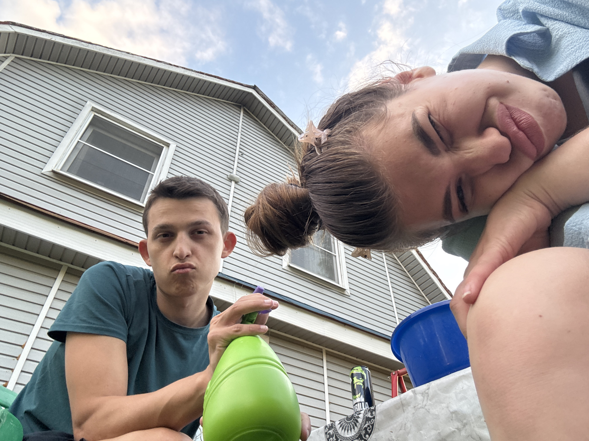 This is a low-angle, wide shot taken outdoors in what appears to be a residential backyard in Meshchery, Russia. The sky is partly cloudy, suggesting it might be daytime. In the foreground, a young man with dark hair and a pouty expression is sitting and holding a bright green spray bottle. He's wearing a dark teal t-shirt. To his right and slightly behind him, a young woman is also in the frame. She's leaning in towards the camera, with her eyes squeezed shut and her lips pursed in a playful grimace. Her hair is pulled up in a bun and adorned with a small, light-colored clip shaped like an animal. She's wearing a light blue garment.
Behind the people, the side of a light gray, vinyl-sided house is visible. It has a white-trimmed window with horizontal blinds. A white roofline with gray siding is visible at the top of the house. A blue plastic tub and a can of what appears to be "Gorilla" energy drink are on a white, patterned surface in the middle ground. The overall impression is of a casual, possibly playful moment captured between two individuals on a mild day.