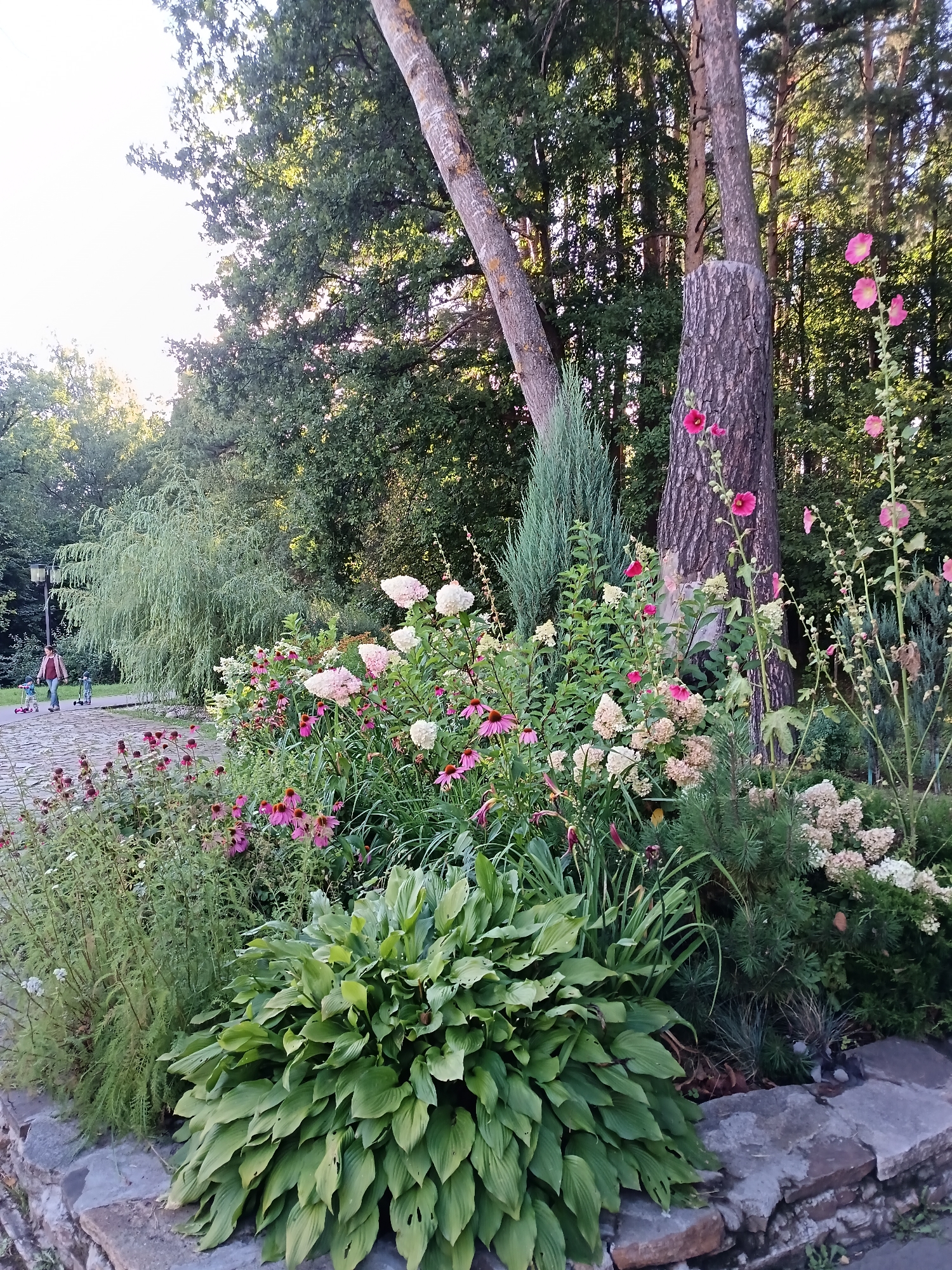 An outdoor daytime scene depicts a stone-lined garden bed filled with lush green foliage and blooming pink and white flowers. In the background, a paved pathway winds through a park-like setting with mature trees. Two children on scooters and an adult are walking on the pathway, with a weeping willow tree to the left and a dense forest in the distance. The weather appears to be overcast.