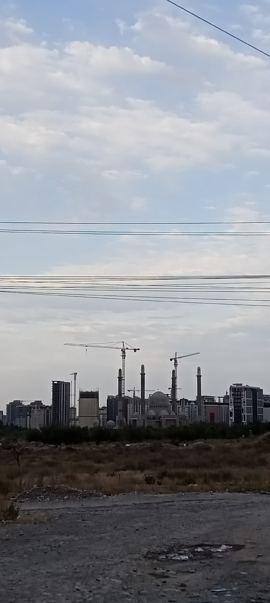 The image depicts a construction site in Bishkek, Kyrgyzstan, with several buildings under various stages of completion. Prominent are multiple tall construction cranes, suggesting active development. The foreground shows a rough, uneven terrain with dry vegetation and some debris. In the mid-ground, a cluster of buildings, including what appears to be a mosque with minarets and a dome, is visible amidst other multi-story structures. Power lines are strung across the sky, contributing to the urban setting. The sky is partly cloudy, with a mix of blue sky and white clouds. The overall scene captures a developing area within the city.