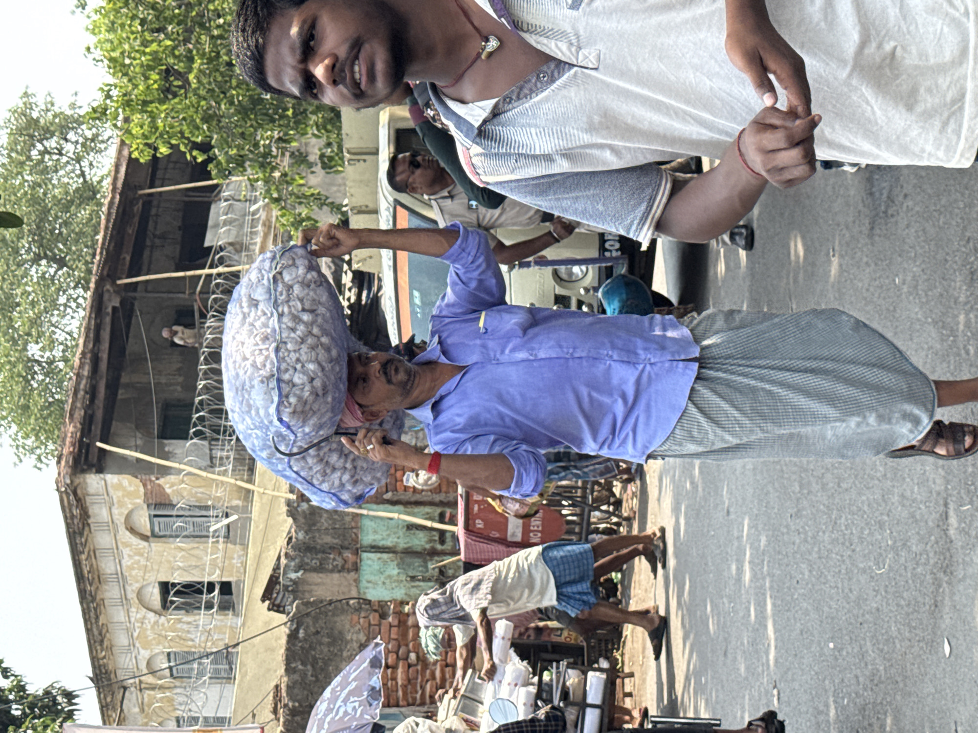 A street scene in Kolkata, India, shows a man carrying a large bag of what appear to be betel nuts on his head. He is wearing a blue shirt and a lungi (a wrap-around skirt) and is walking along a road. Other people are nearby, including street vendors with carts and a man in the foreground. A building with visible signs of age is located in the background.