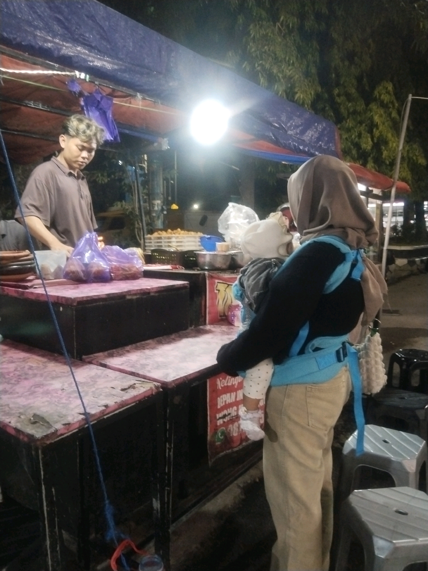 In an outdoor nighttime scene at a street food stall, likely in Pemalang, Indonesia, a young man with styled, light-colored hair stands behind a food cart. He is wearing a brown polo shirt and appears to be attending to the cart. In front of him, on the cart's counter, are several purple plastic bags filled with food items.
To the right, a person wearing a light brown hijab and a dark top, also in khaki pants, is holding a baby. The baby is wrapped in a light-colored outfit and is secured in a light blue baby carrier. The food cart has a pink and white checkered countertop and dark bases. Various items, including plates and containers, are visible on and around the cart. In the background, trees are visible, and the lighting suggests an urban or semi-urban setting. There are also plastic stools in the foreground. A sign with Indonesian text is partially visible on the cart.