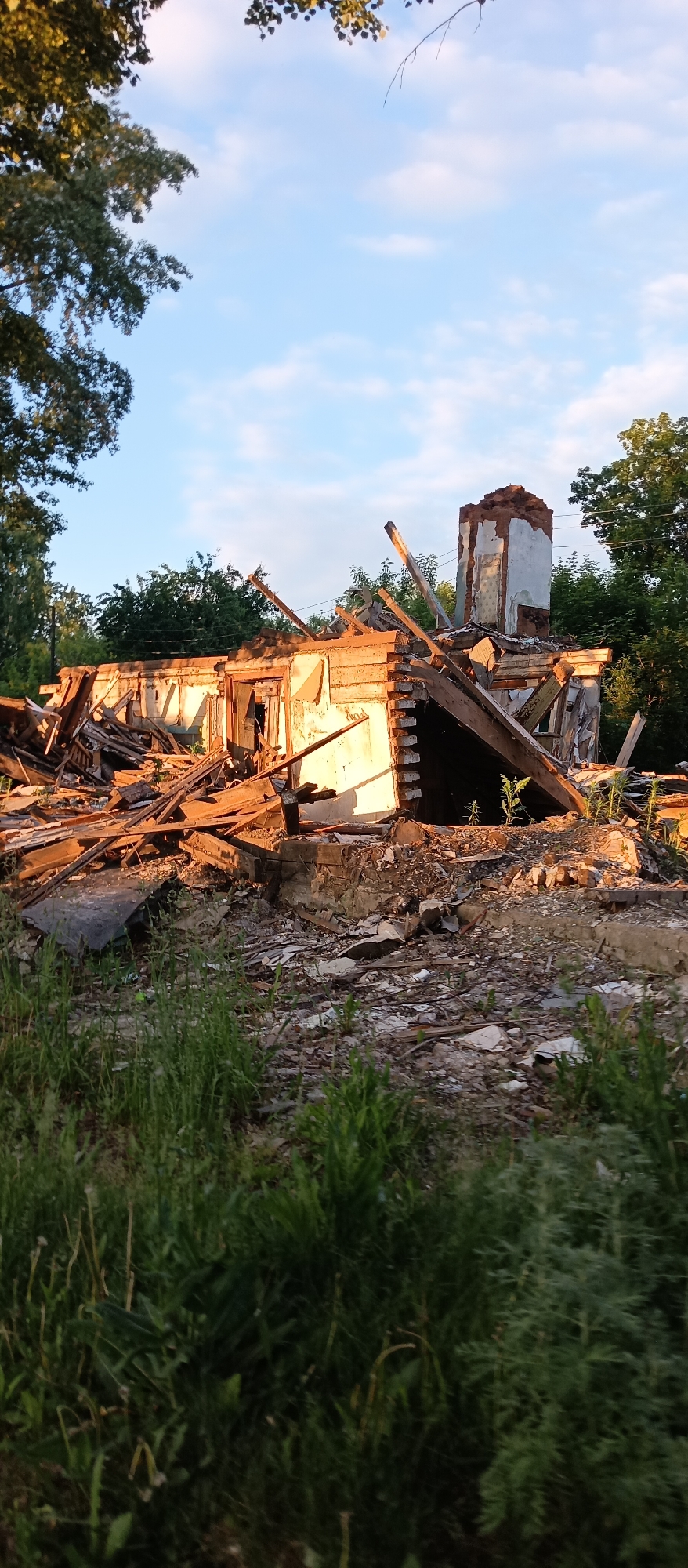 This image, captured in the city of Chaikovsky, Russia, depicts a partially demolished building structure under a clear sky with scattered white clouds. The scene is illuminated by warm, golden-hour light, casting long shadows and highlighting the texture of the debris.
The central subject is a ruined building, primarily composed of what appears to be wooden framing, white-painted or plastered exterior walls, and brickwork. The structure is significantly collapsed, with large quantities of wooden beams, planks, and other building materials forming a substantial pile of rubble in the mid-ground. One section of an exterior wall, painted a light color, remains partially standing, featuring a dark, open doorway. To the right and rear of this wall, a prominent, tall, and narrow brick chimney stands, heavily damaged with exposed red brick and remnants of white plaster. Several large wooden structural elements are angled precariously within the debris pile.
The foreground is characterized by dense green grass and weeds, partially obscuring the lower portions of the rubble. Beyond the ruined building, a line of mature green trees forms the background. No individuals, animals, or active machinery are visible in the scene, suggesting a state of abandonment or paused demolition.