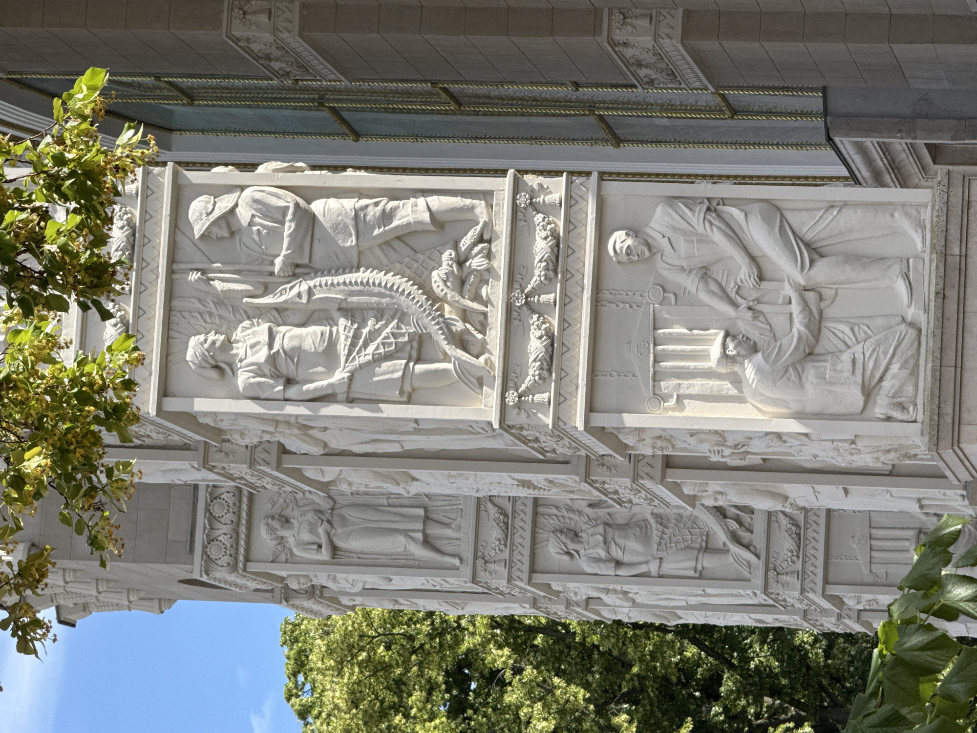 The image shows a detail of the bas-relief frieze on the exterior of the State Kremlin Palace in Moscow, Russia. The frieze is made of white stone and depicts various scenes. In the foreground, a column of the palace is visible, adorned with carved figures. One section of the frieze shows two people, a man and a woman, interacting with a large, scaly creature. Another section depicts two men engaged in what appears to be a craft or industrial activity, with one standing and the other kneeling. The background features greenery from trees and a bright blue sky. The architectural style is grand and classical, typical of Soviet-era monumental art.