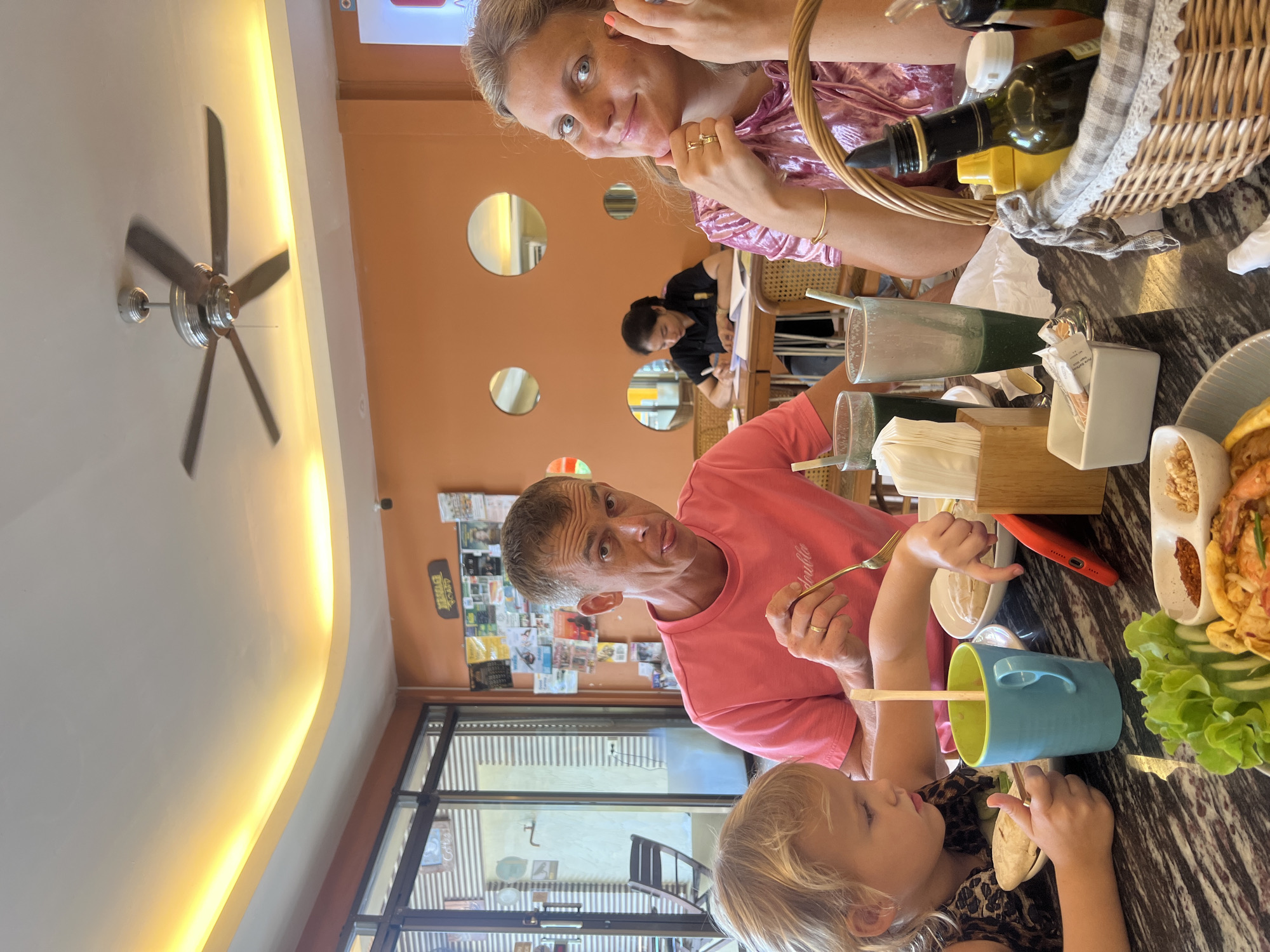 The image captures a family dining at a restaurant in Koh Phangan, Thailand. The scene is set at a table with a dark, marbled surface. On the table, there are plates of food, including what appears to be Pad Thai with shrimp and lettuce, as well as drinks in tall glasses and a blue mug. A woven basket filled with condiments such as bottles of sauce and mustard is also visible.
In the foreground, a young blonde girl with a patterned top is eating, holding a spoon. To her right, a man in a pink t-shirt is also eating and looking towards the camera with his tongue sticking out. To the far right, a blonde woman with a pink, shimmering top is smiling and touching her face. In the background, a woman of East Asian descent is seated at a table, writing in a notebook.
The restaurant's interior is characterized by orange walls adorned with decorative circular cutouts and various posters and images. A ceiling fan is visible above, and strip lighting illuminates the ceiling. Large windows with blinds are present on the left side of the image, revealing a glimpse of an outdoor seating area. The overall atmosphere suggests a casual, familial meal in a tropical setting.