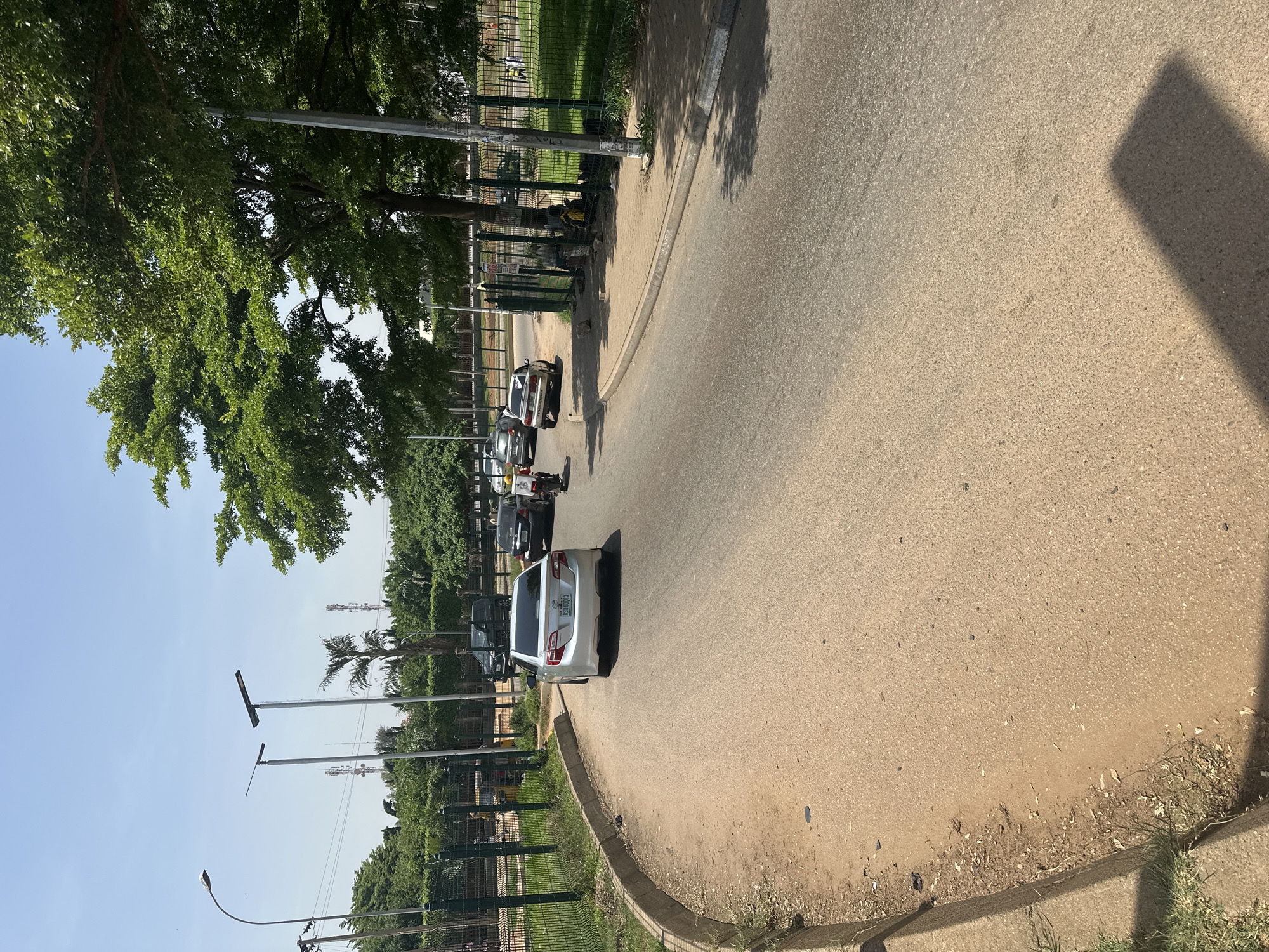 The image captures a street scene in Abuja, Nigeria, on a clear, sunny day. A light-colored sedan, identified by its license plate as R589DUQ, is parked on the right side of a paved road that curves to the left. The car appears to be a Toyota Camry. In front of the sedan, a motorcycle is visible, carrying a person in a red jacket and white helmet, and a delivery box. Further up the road, other vehicles, including a dark SUV and a few other cars, are driving or parked. The road is bordered by a sandy shoulder on the left and a green metal fence on the right, behind which a grassy area and trees are visible. Tall light poles with solar panels are positioned along the road. In the background, several telecommunications towers can be seen against the blue sky. The overall environment suggests an urban setting with infrastructure for public services and transportation.