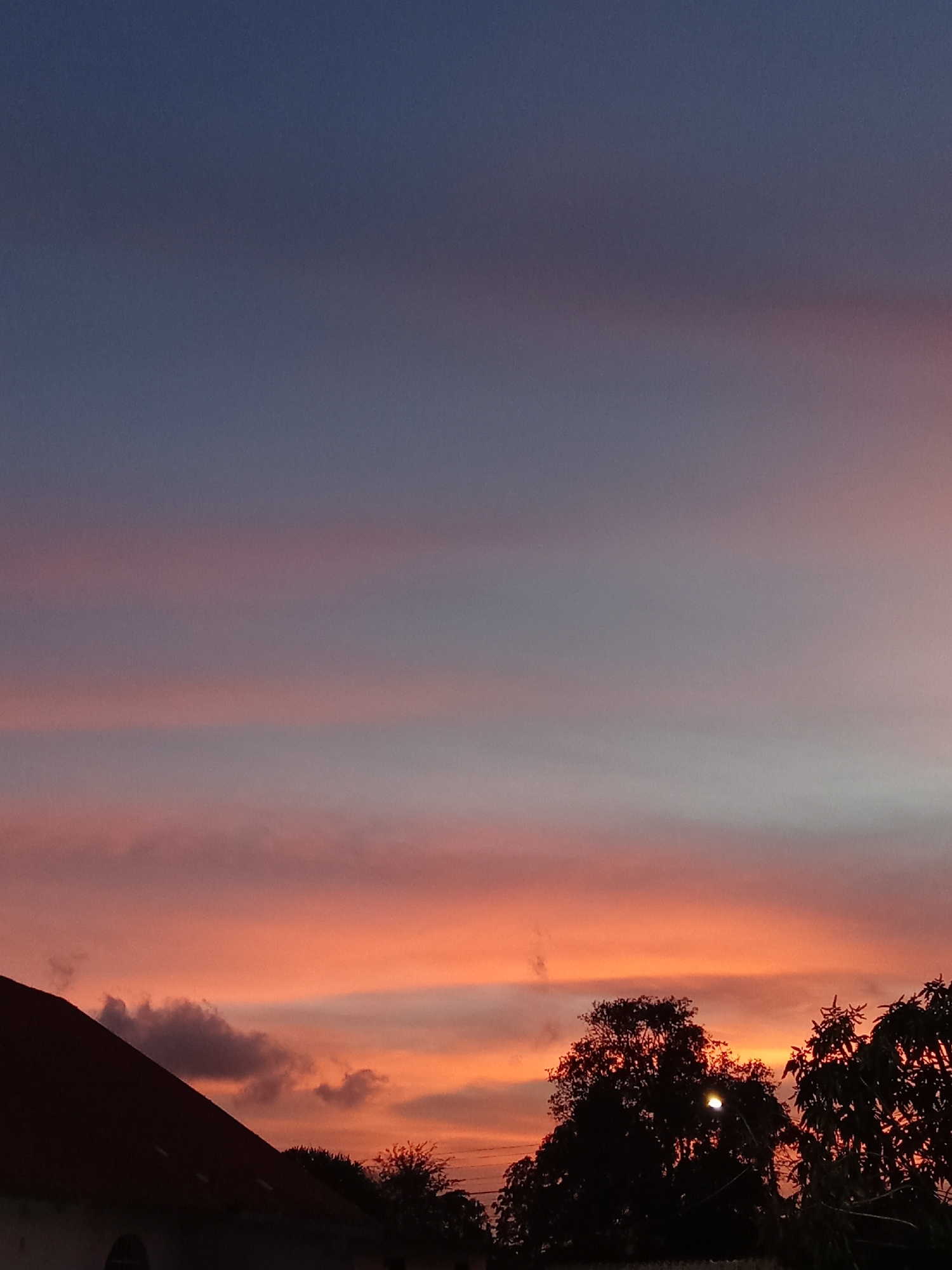 This image depicts an outdoor scene in Juan Griego, Venezuela, at twilight, likely during sunset. The sky occupies the majority of the frame, displaying a distinct color gradient. The upper portion of the sky is a deep, dark blue, which transitions downwards into horizontal bands of muted purple and pink clouds. Below these, the sky brightens significantly to vivid orange and red hues, indicating the position of the setting sun. Scattered, darker cloud formations are visible, particularly in the mid-section and near the horizon. In the lower left foreground, a section of a dark, reddish-brown pitched roof is visible in silhouette, belonging to an unseen structure. In the lower right and center, dense tree canopies are also silhouetted against the illuminated sky. A single, distinct point of artificial light, consistent with a streetlamp or similar fixture, is visible embedded within the foliage of one of the central trees. No individuals or specific activities are present.