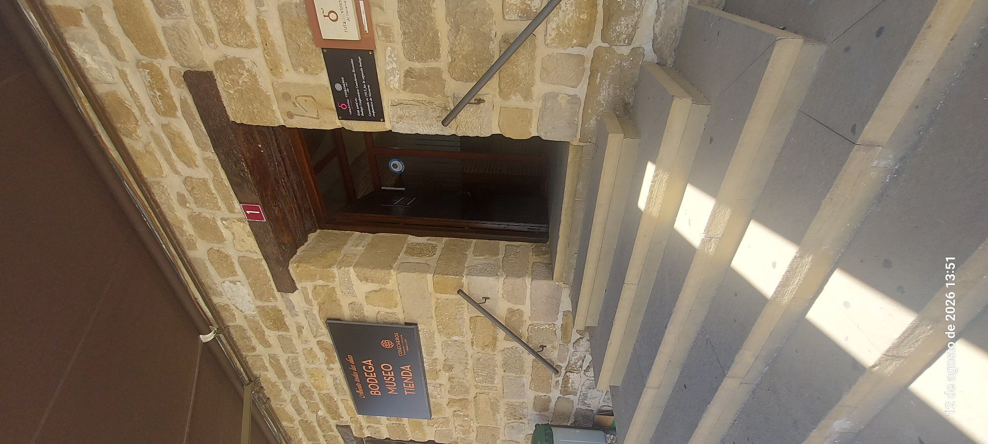 The image shows the exterior of a building constructed of light-colored stone blocks. A dark brown wooden door is set within a stone archway. To the left of the doorway, a dark rectangular sign is mounted on the stone wall. The sign has white text that reads "Abierto todas los dias", "BODEGA", "MUSEO", and "TIENDA", with a circular logo containing text below. Above the doorway, a small red square sign with the number "1" in white is visible. To the right of the doorway, another dark sign is mounted on the wall, featuring a red symbol and text that includes "ruta del vino de Navarra".
In front of the building are a series of wide, light-colored stone steps leading up to the entrance. Metal handrails are attached to the stone wall on either side of the steps. Sunlight casts distinct shadows across the steps. To the left of the steps, near the bottom, a green cylindrical object, possibly a trash receptacle, is visible, along with a brown cardboard box. The overall environment suggests an entrance to a public establishment, likely a winery or cultural center, in Olite, Spain. A timestamp at the bottom of the image indicates "12 de agosto de 2026 13:51".