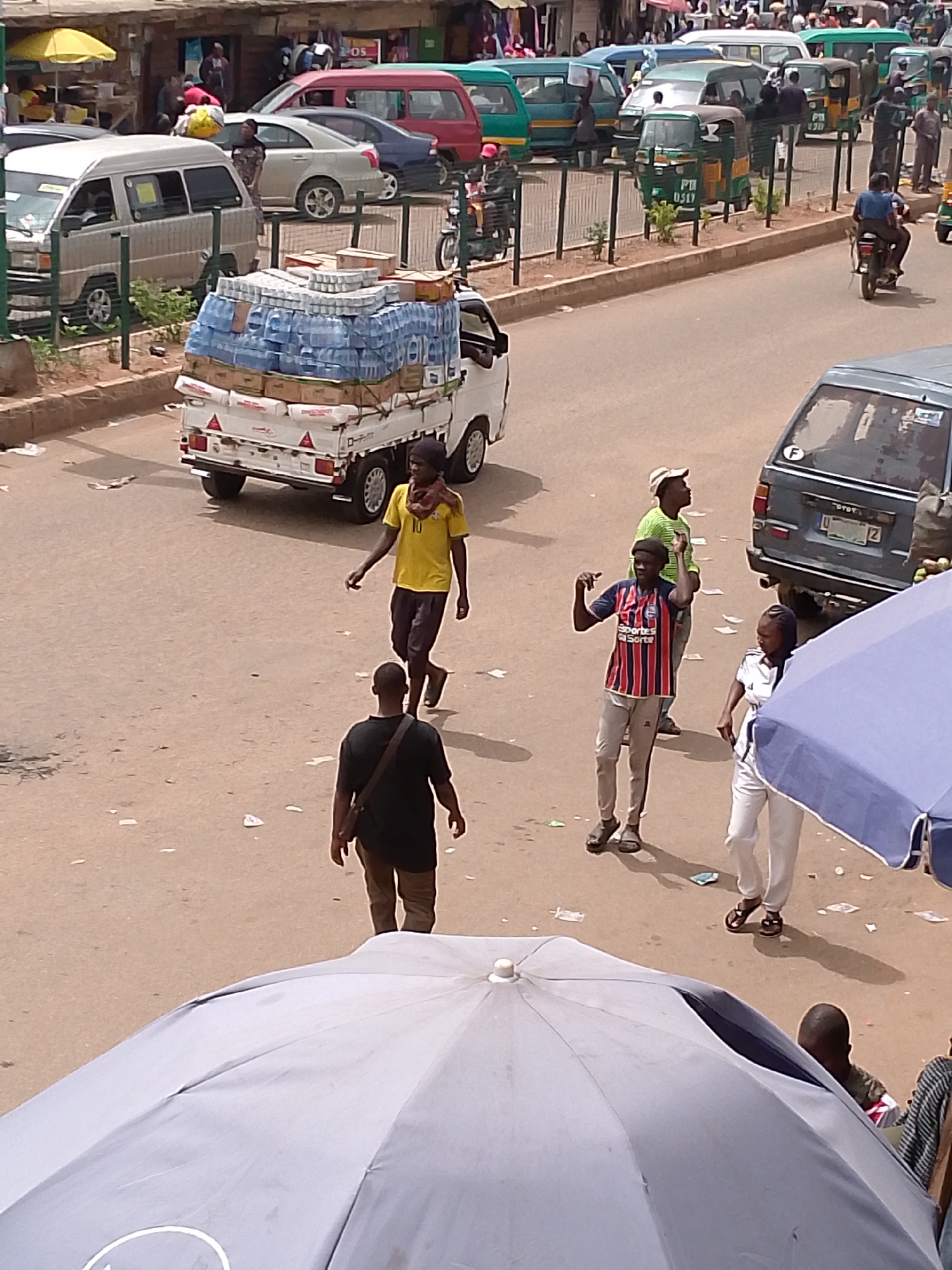 The image depicts a street scene in an urban area, possibly in Africa, based on the appearance of the people and vehicles. A small white truck loaded with a large stack of bottled water is prominent in the foreground. Several people are walking on the street, including a man in a yellow shirt, a man in a black shirt, a man in a striped soccer jersey, and a woman in white clothing. In the background, there is a line of parked vehicles, including small vans, cars, and auto-rickshaws (tuk-tuks). There are scattered pieces of litter on the street. The view is taken from an elevated position, possibly from a building or upper story. A large blue umbrella is partially visible in the lower part of the frame.