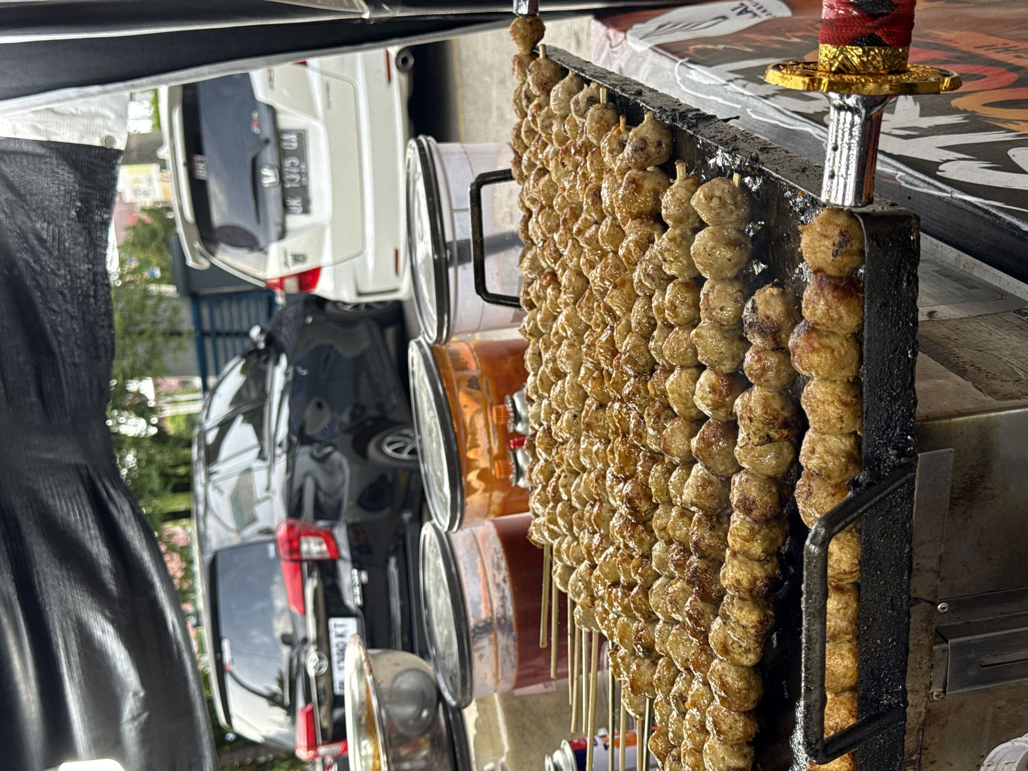 A close-up, eye-level outdoor shot focuses on a food stall in Lhokseumawe, Indonesia, featuring rows of grilled meatballs on skewers cooking on a flat-top grill. The meatballs are golden brown and glistening with sauce, some showing char marks. Behind the grill, two large plastic containers hold red sauce and a lighter-colored sauce or condiment. In the background, several cars are parked, including a black SUV on the left and a white hatchback on the right. The stall is covered by a black tarp overhead. The lower right corner of the image shows a decorative, sword-like handle, possibly part of the stall's branding or equipment. The scene appears to be a street food vendor setup, likely operating in an urban or semi-urban area of Lhokseumawe. There are no people visible in the frame.