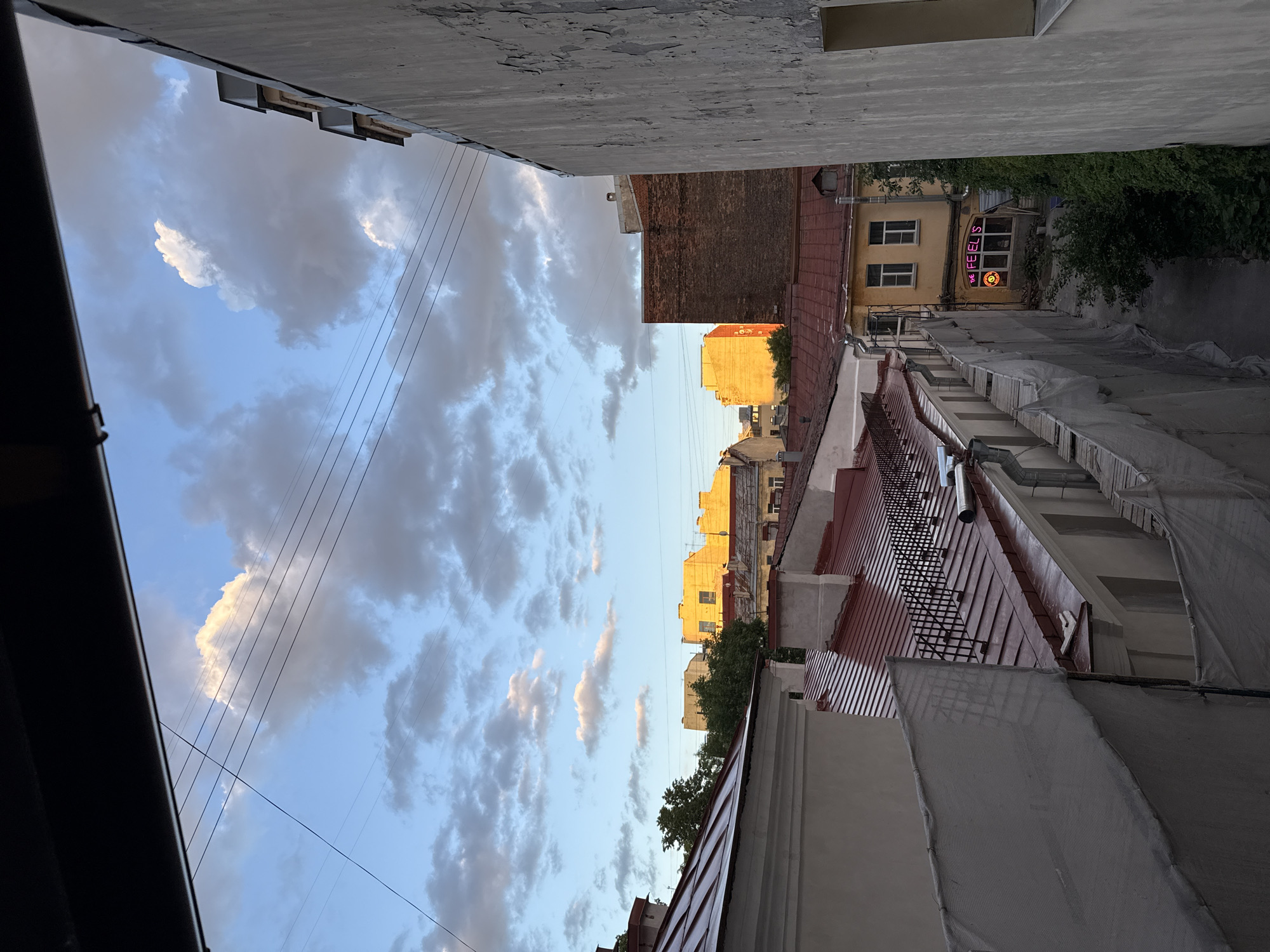 The image depicts a view of Saint Petersburg, Russia, from a building's upper level, looking out over rooftops and into a courtyard. The scene captures the late afternoon or early evening sky, with dramatic clouds illuminated by the setting sun, casting a warm glow on several buildings.
In the foreground, to the right, is a weathered concrete wall with peeling paint, suggesting an older structure. A dark overhang forms the top edge of the frame, implying the viewpoint is from an interior space looking outwards.
Below the sky, a series of red-tiled roofs dominate the mid-ground. Some have visible metal railings along their edges, and chimney stacks are present. Construction netting or tarps are visible in the lower left, draped over scaffolding or structures, indicating ongoing renovation or building work.
Further back, buildings of various architectural styles and colors are visible. Notably, a block of buildings is bathed in a strong, orange sunlight, highlighting their yellow facades and windows. To the left of this, trees are interspersed among the structures.
In the courtyard area, visible between buildings and behind the construction netting, a yellow building with two windows has a neon sign in pink letters that reads "THE FEEL'S" with a circular graphic resembling an emoji or a smiley face. This suggests a commercial establishment, possibly a bar or cafe, within the courtyard.
The overall impression is an urban landscape, characteristic of historic European cities, with a mix of old and new construction, residential and potentially commercial spaces, and the ever-present sky as a backdrop. The lighting suggests a specific time of day, adding a dramatic quality to the urban panorama.