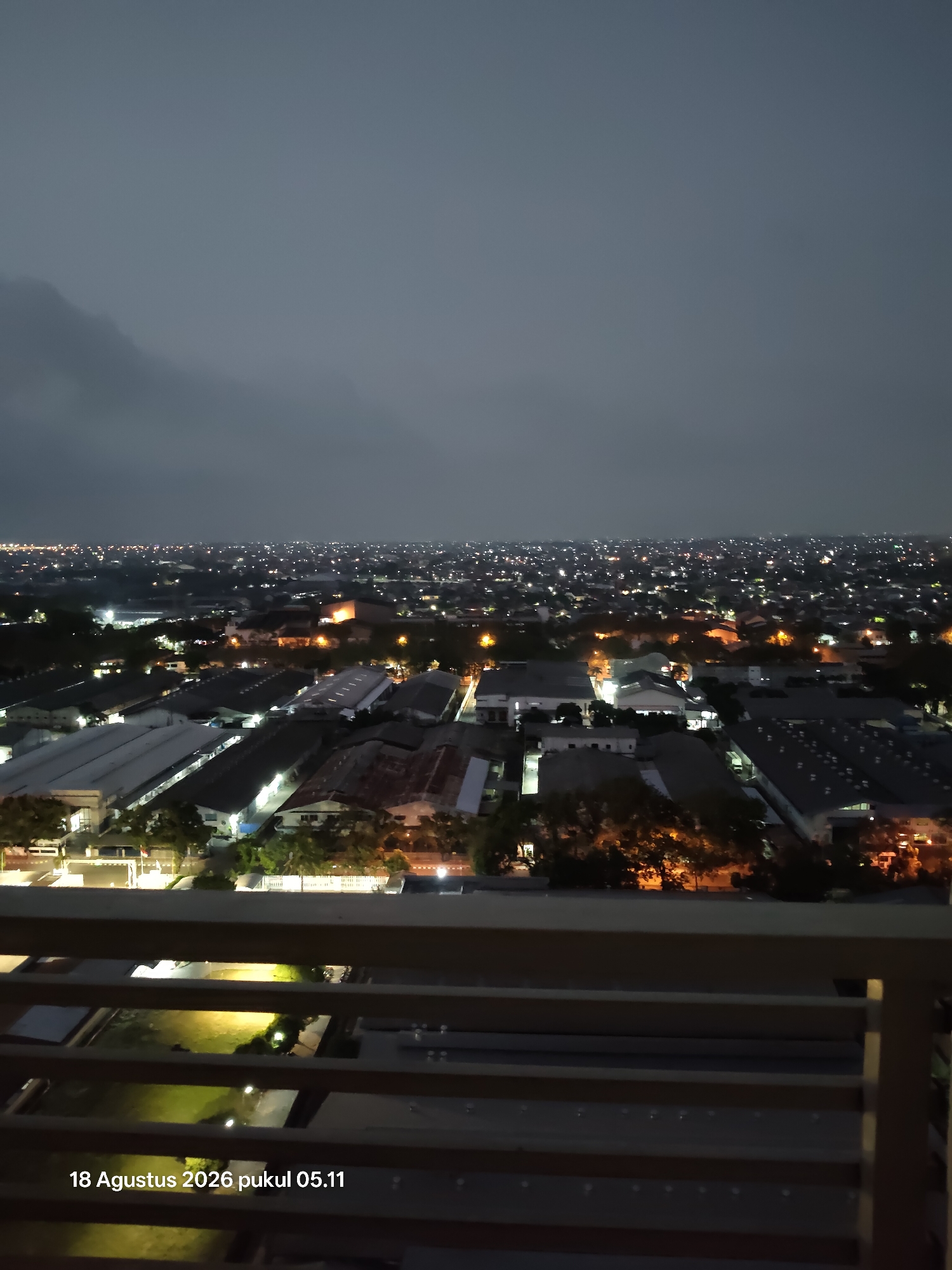 The image depicts a nighttime view of an urban landscape, likely industrial or commercial areas, from an elevated vantage point. The sky is dark and overcast, with a faint glow on the horizon indicating distant city lights. The foreground features a balcony railing, suggesting the viewpoint is from a building. Below, a sprawling city unfolds, characterized by numerous buildings with illuminated windows and streetlights. The architecture appears to be a mix of industrial warehouses with large, flat roofs and residential structures. Trees are scattered throughout the scene, some illuminated by artificial lights. The overall impression is one of a busy urban environment at the start of the day, as indicated by the timestamp "18 Agustus 2026 pukul 05.11" which translates to "August 18, 2026 at 05:11 AM". The location is specified as Surabaya, Indonesia.